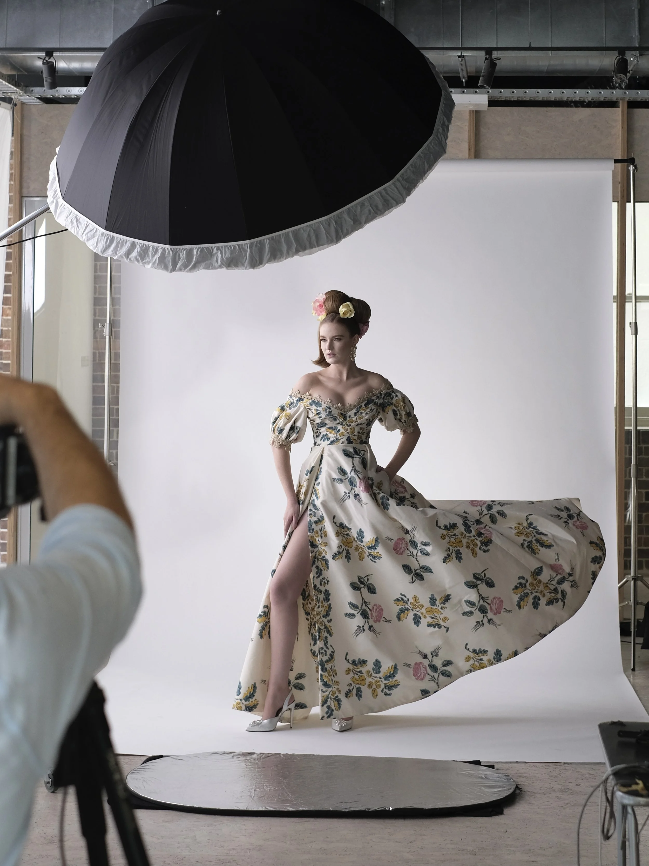 A woman modeling a floral off-shoulder gown with a high slit, standing in front of a white backdrop during a photoshoot with studio lighting equipment.