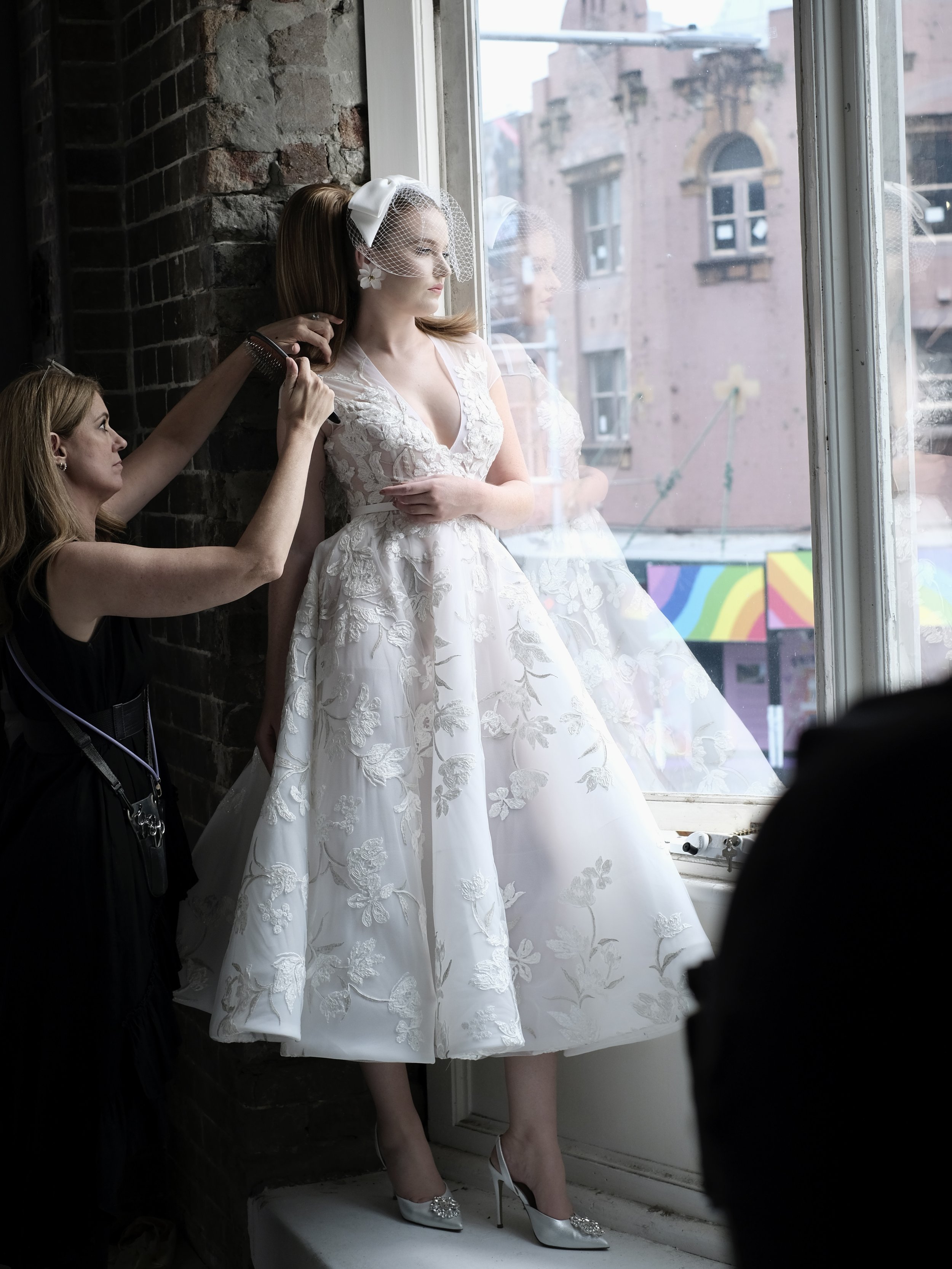 A bride in a white lace wedding dress with floral accents sits on a window ledge, while a stylist adjusts her hair. The bride wears a birdcage veil and white high heels with embellishments, inside a brick-walled studio with large windows showing buil