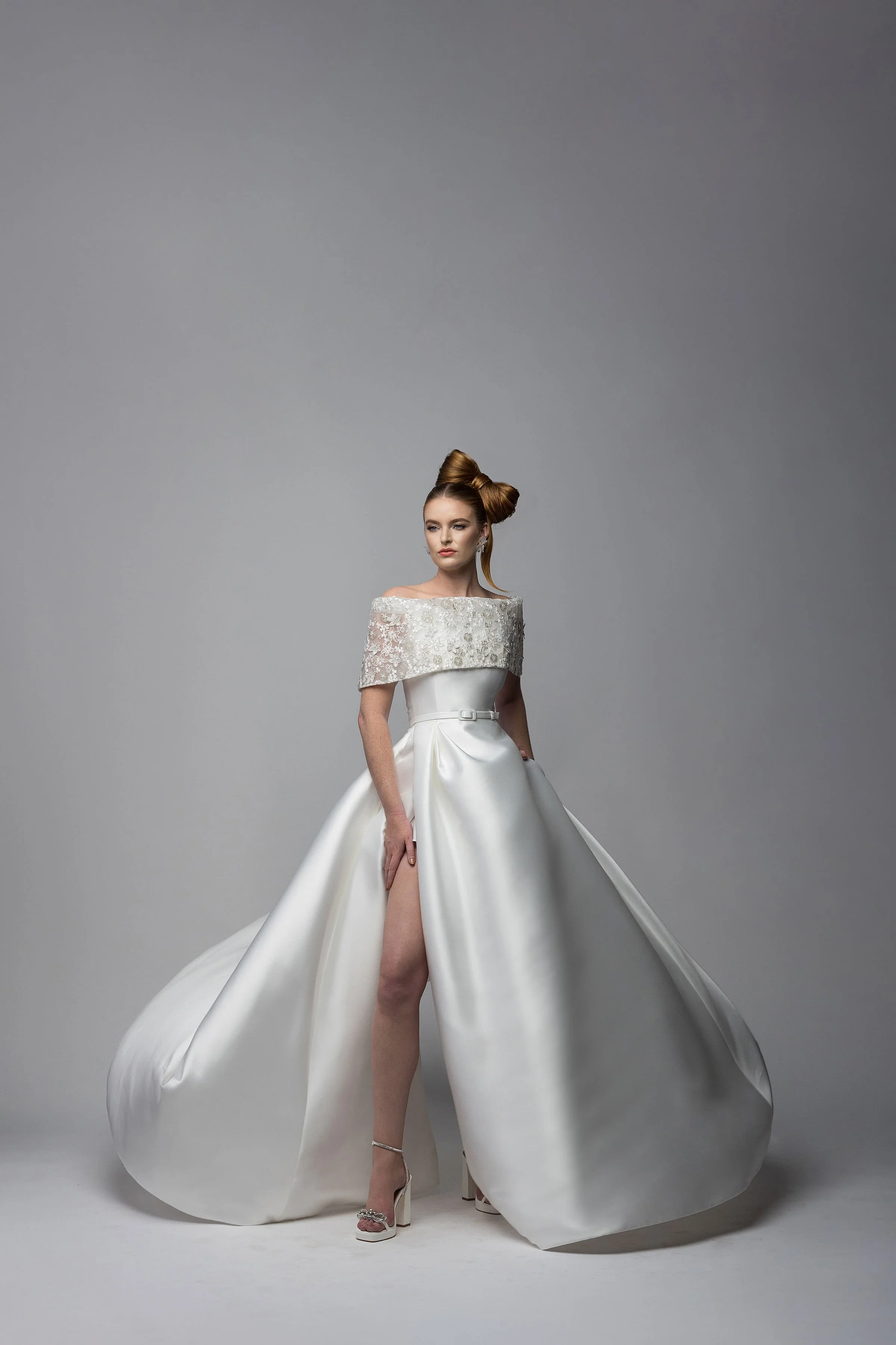 A woman in a white wedding dress with an off-the-shoulder lace bodice and a full satin skirt, standing against a plain gray background.