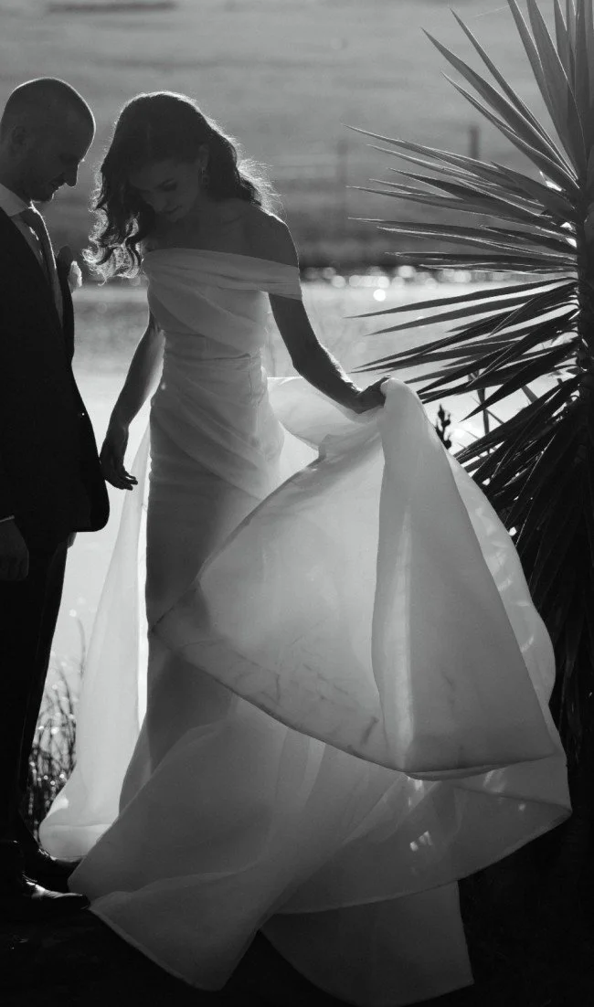 A black and white photo of a bride and groom standing outdoors near water, with the bride holding her wedding dress and gazing down, and the groom looking at her, beside a palm tree.