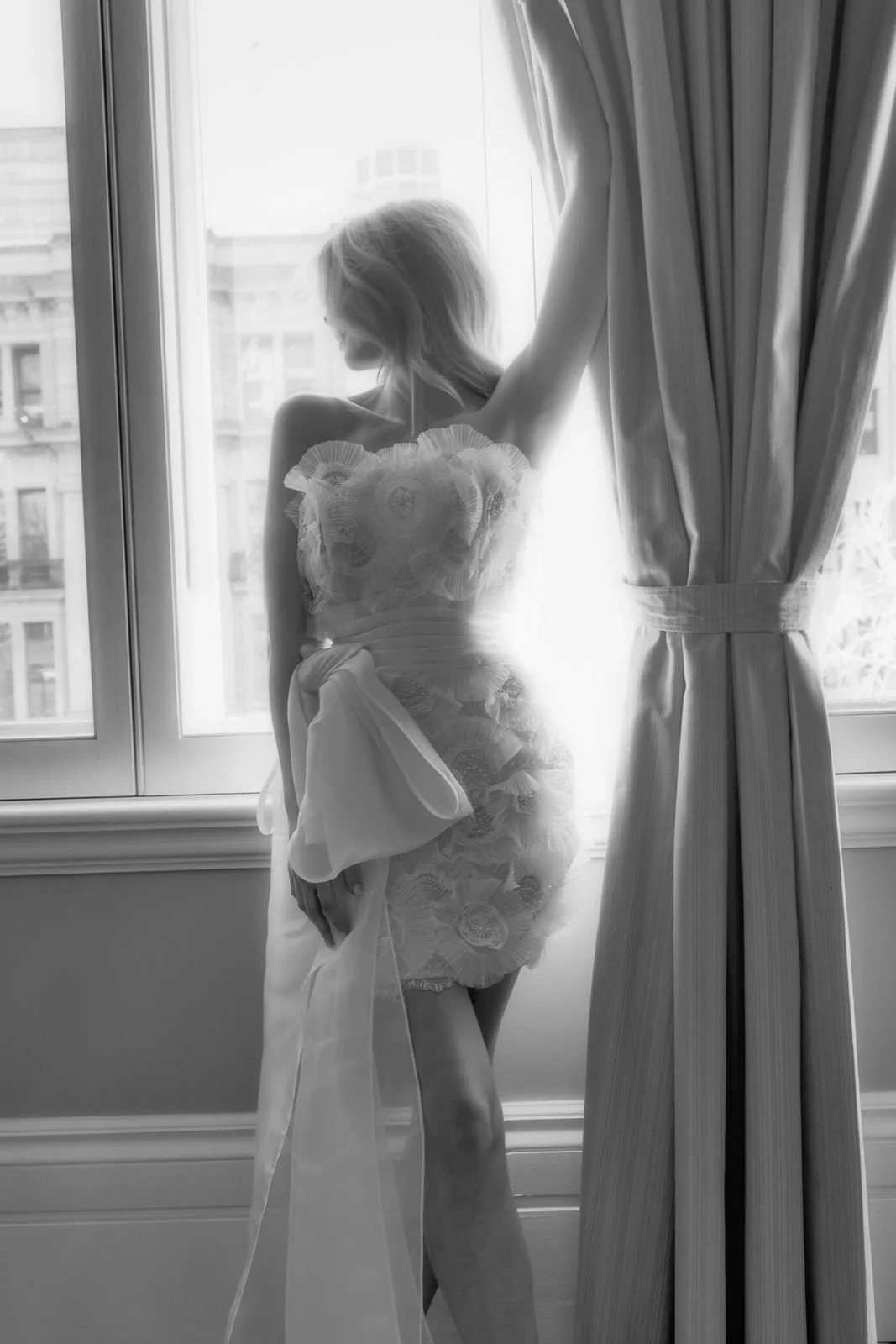 A woman standing by a window, holding back curtains and looking outside, dressed in a sheer lace dress with floral patterns, with soft backlighting highlighting her silhouette.