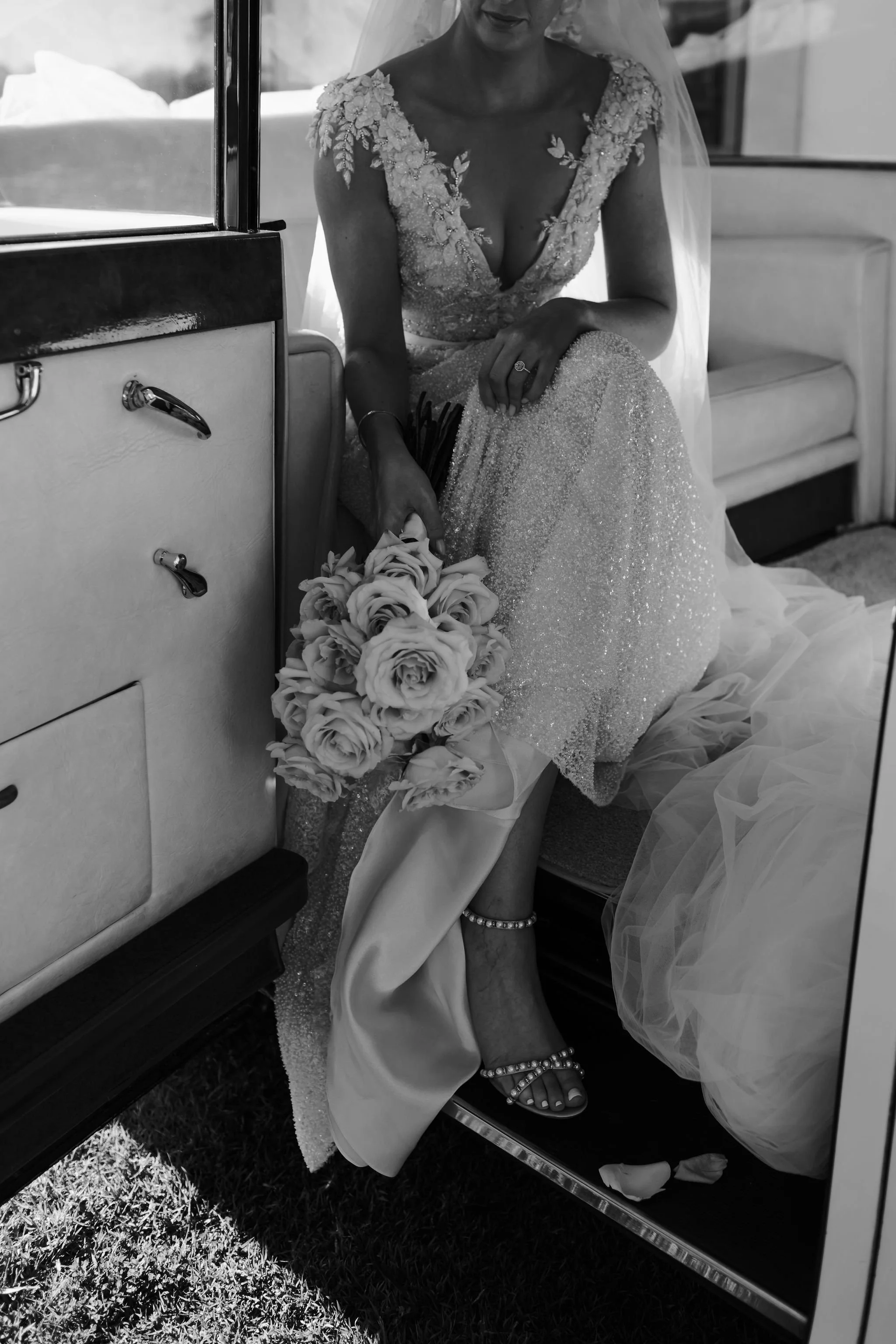 A bride in a wedding dress holding a bouquet of roses sits inside a vehicle.