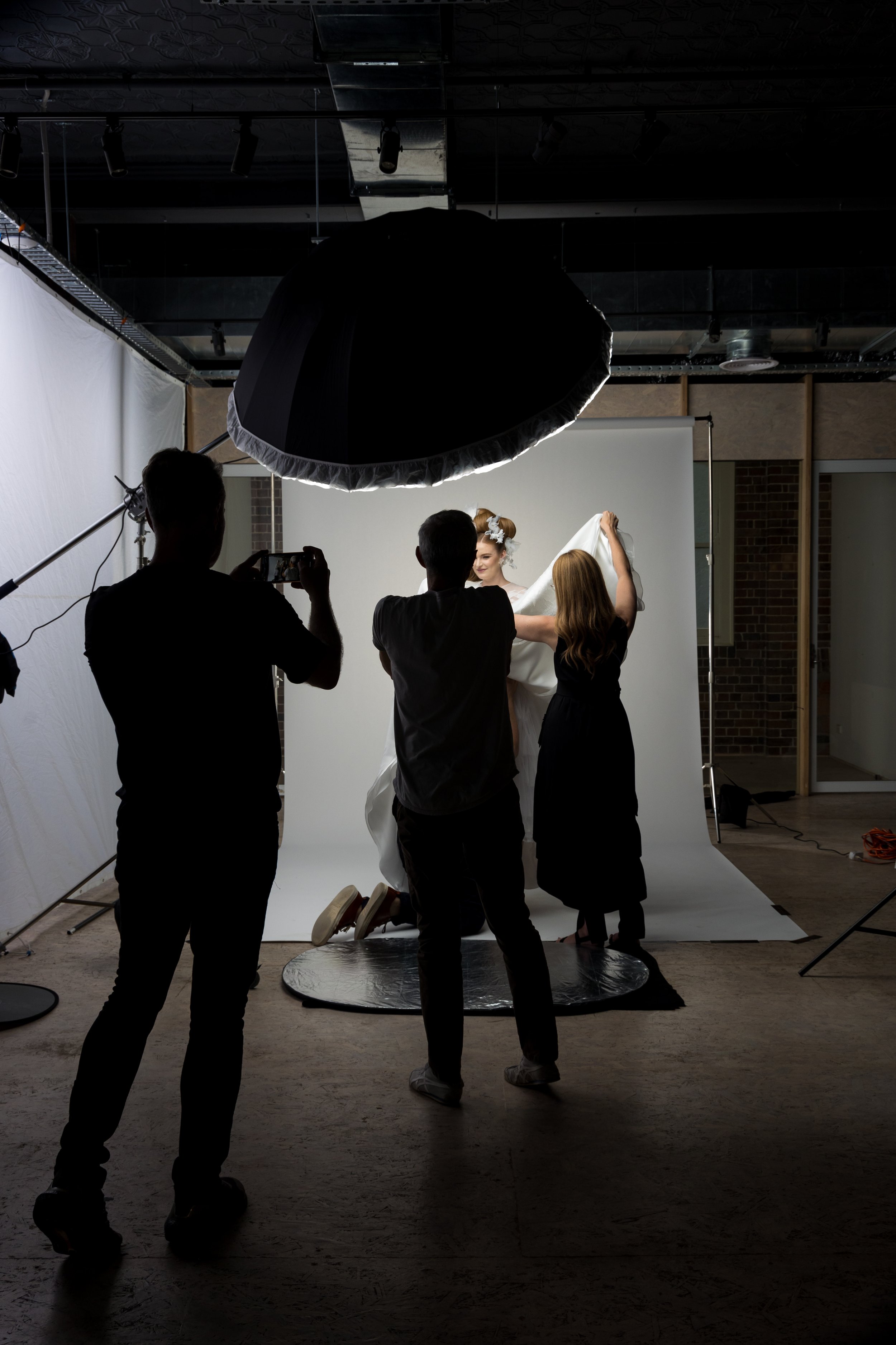 Photographers and assistant capturing a portrait of a woman in a studio with a white backdrop, professional lighting equipment in use.