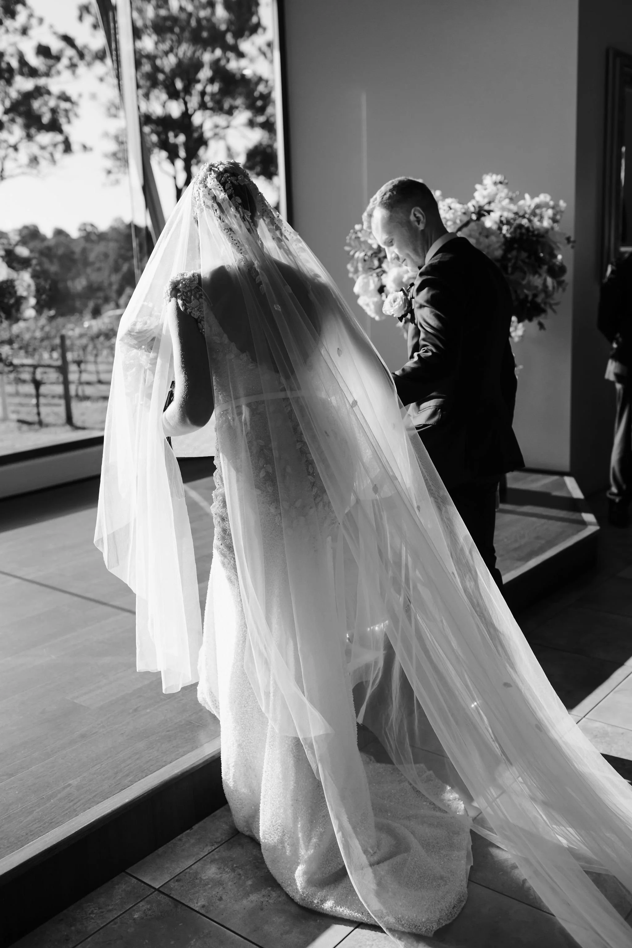 Black and white photo of a bride with a veil and a groom in a tuxedo during a wedding ceremony, standing inside near a large window with sunlight streaming in.