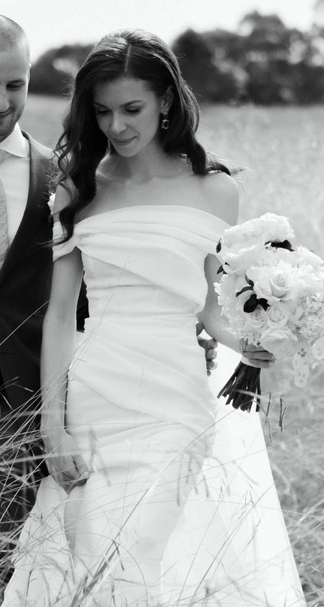 A black and white photo of a bride holding a bouquet of flowers, wearing an off-the-shoulder wedding dress, standing outdoors in a grassy field.