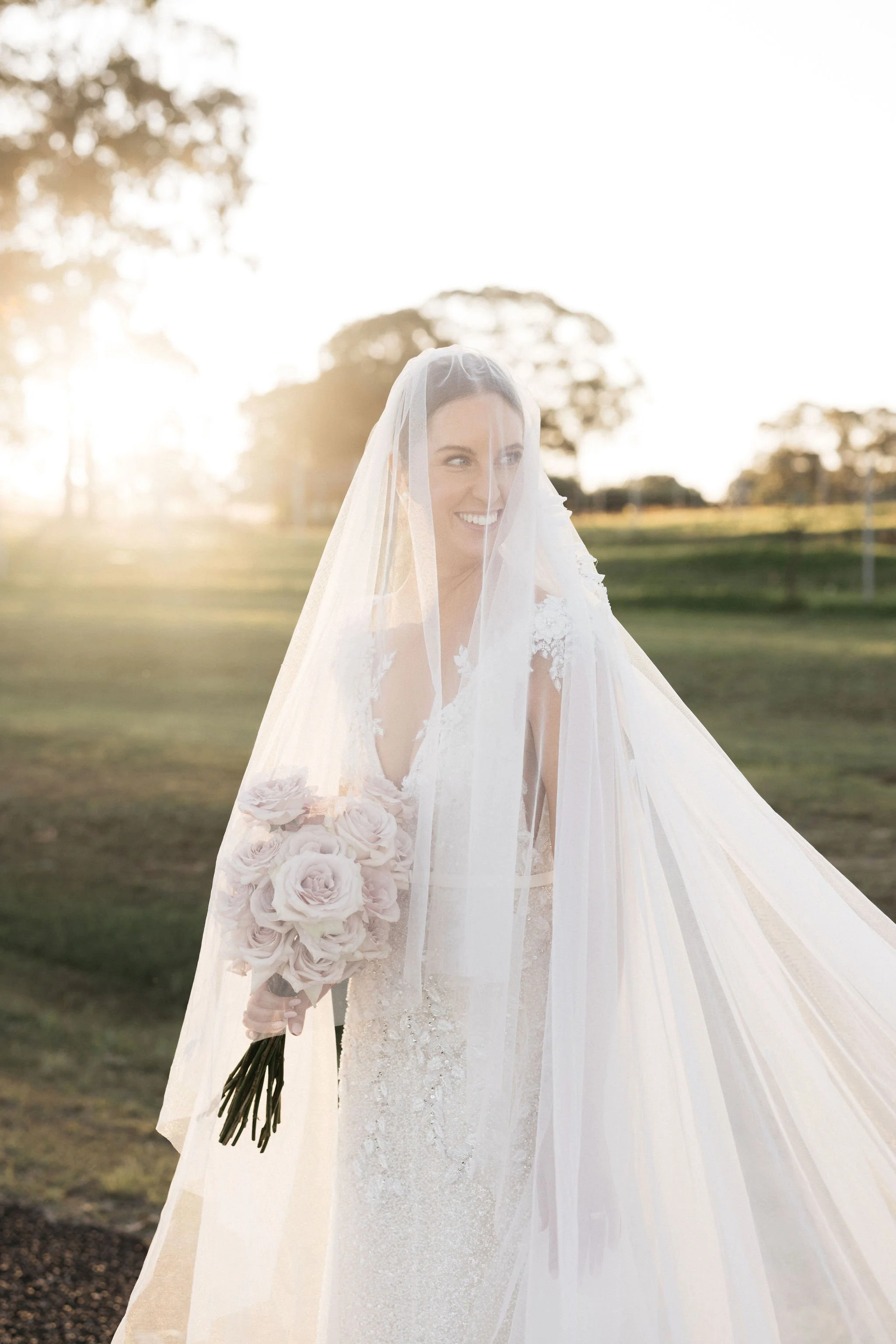 A bride in a white wedding dress and veil holding a bouquet of light pink roses outdoors during sunset, with trees in the background.