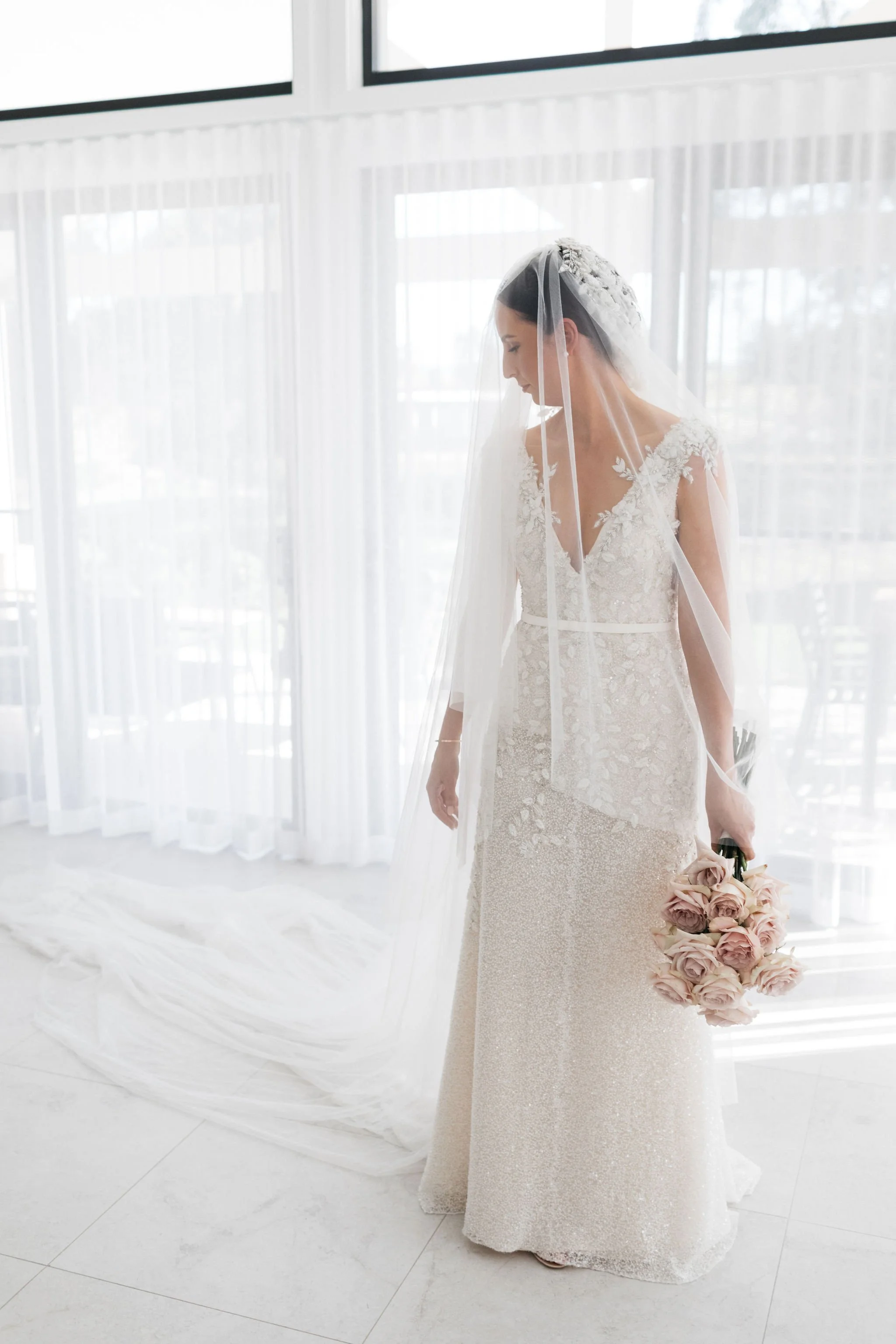 Bride in a white wedding dress with lace details, holding a bouquet of pink roses, standing indoors near large windows with sheer curtains.