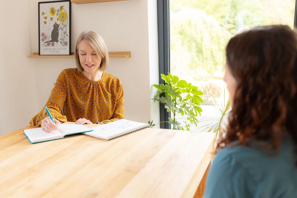 A woman with shoulder-length blonde hair wearing a yellow patterned blouse sitting at a wooden table, writing in a notebook. Another woman with curly brown hair wearing a blue top sits across from her. There is a window with a view of greenery and a plant beside the window. A framed picture of a black cat with yellow flowers hangs on the wall behind the first woman.