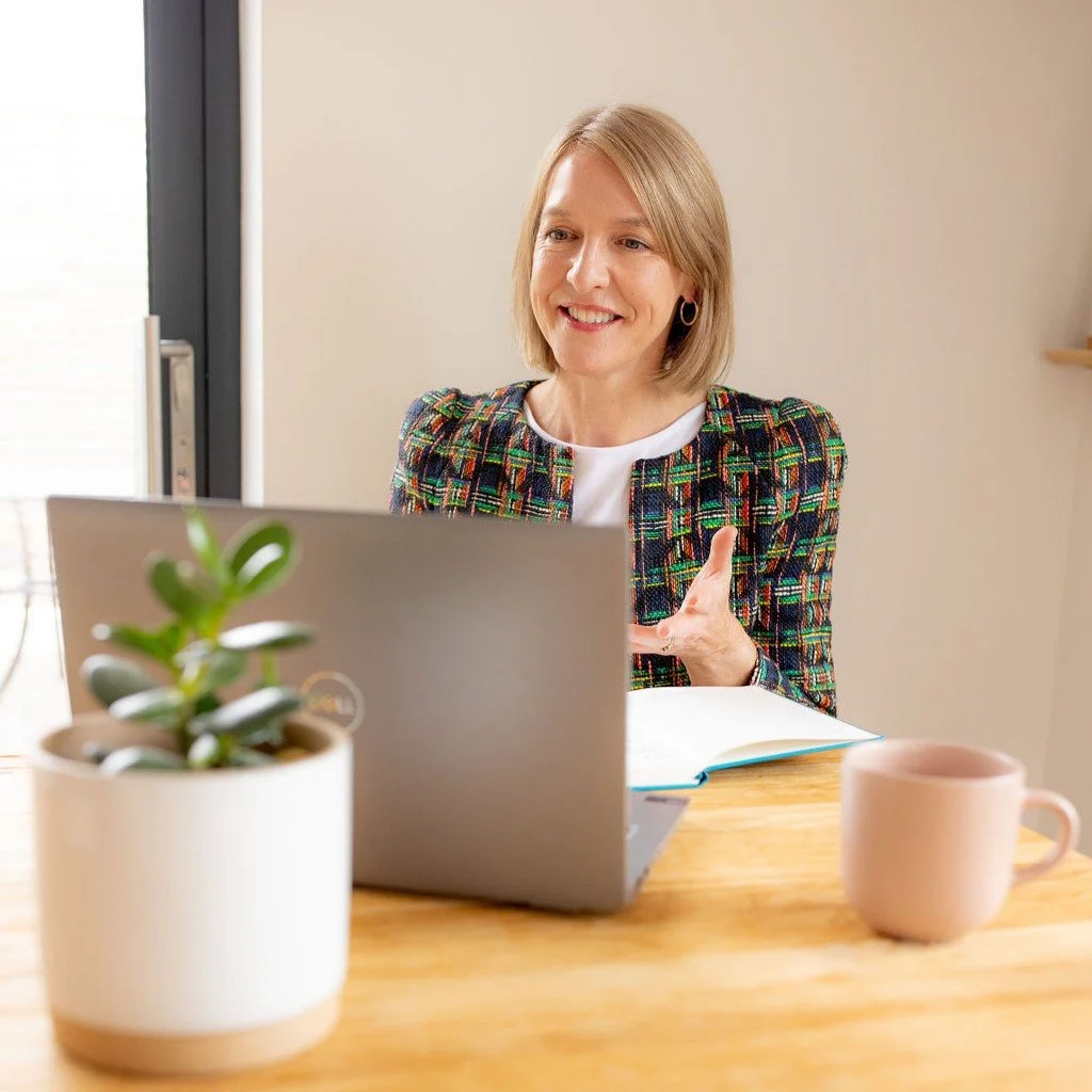 A woman with blonde hair smiling and gesturing while sitting at a wooden table with a laptop, a pink mug, a small potted plant, and an open notebook.