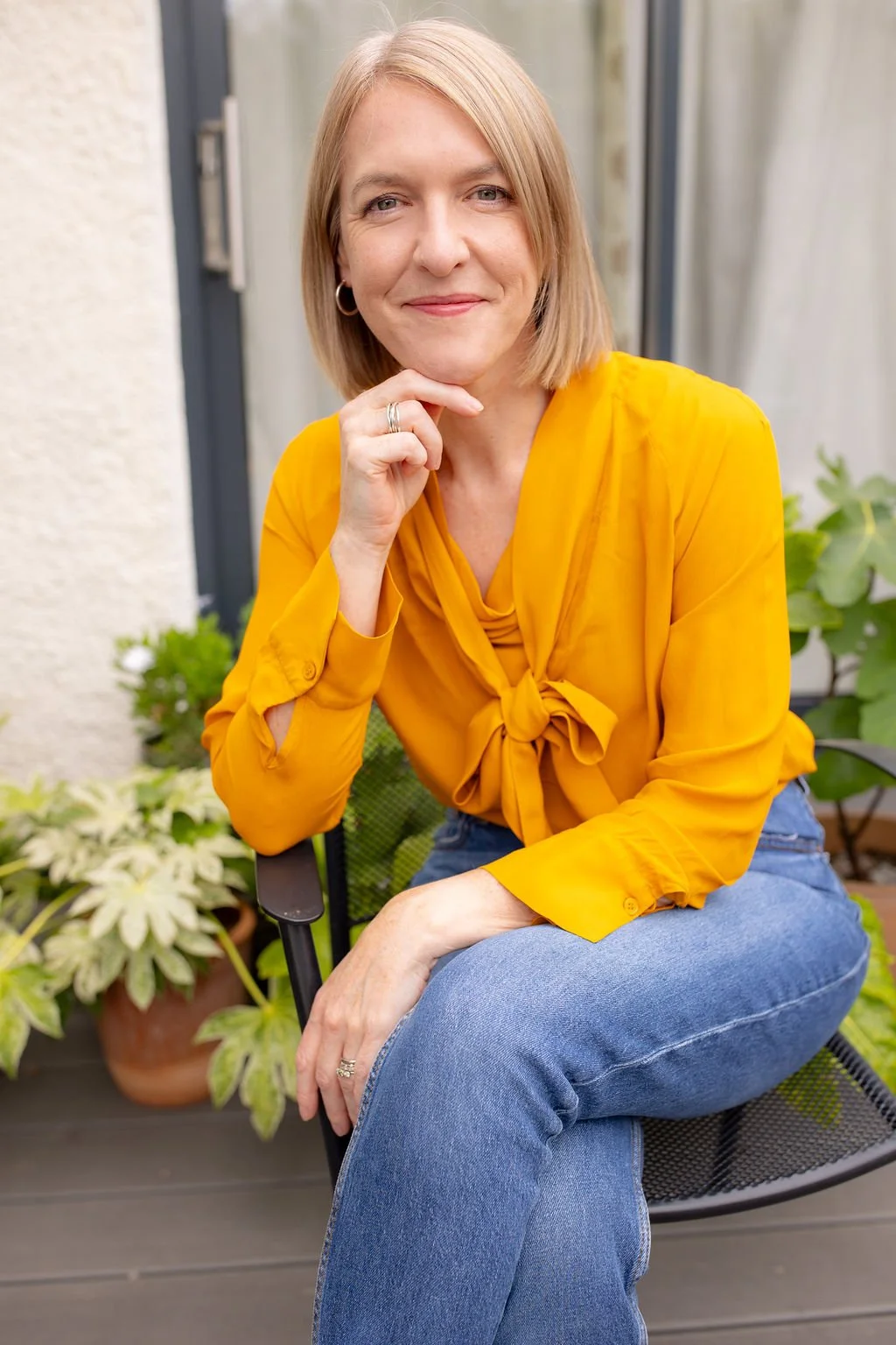 A woman with blonde hair, wearing a yellow blouse and blue jeans, sitting outdoors on a chair with plants in the background, smiling at the camera.