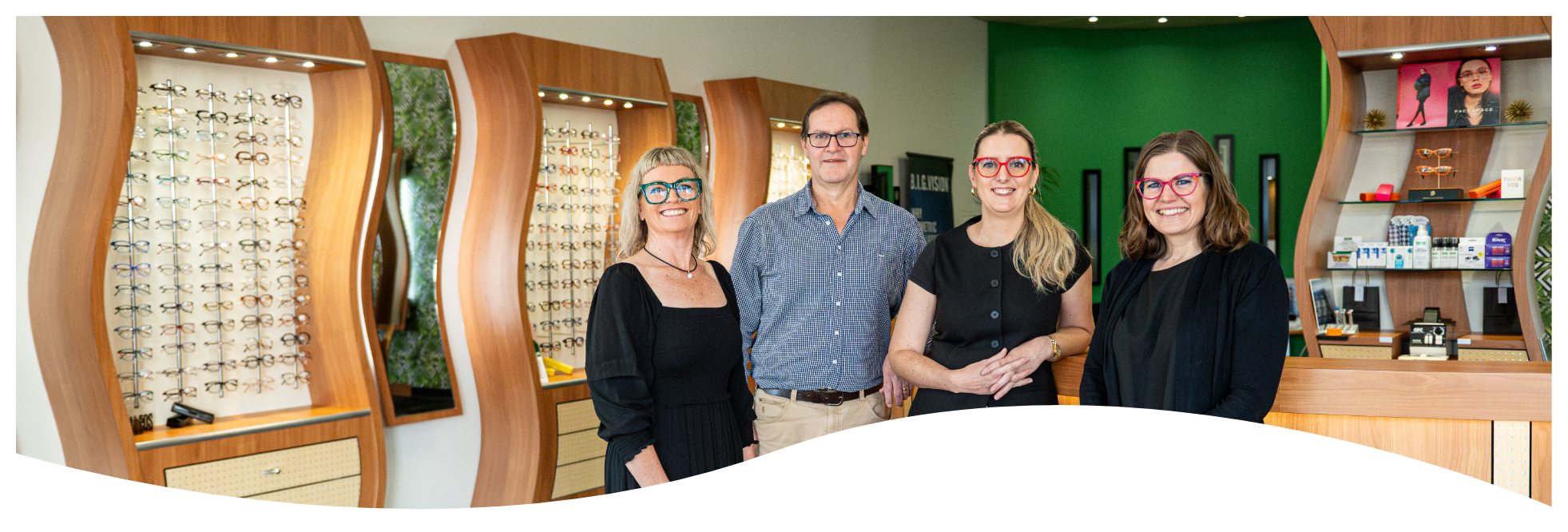 Four people, three women and one man, standing together inside an eyeglasses store, smiling at the camera. The store displays many eyeglasses on wooden shelves behind them, with a green wall and framed pictures in the background.