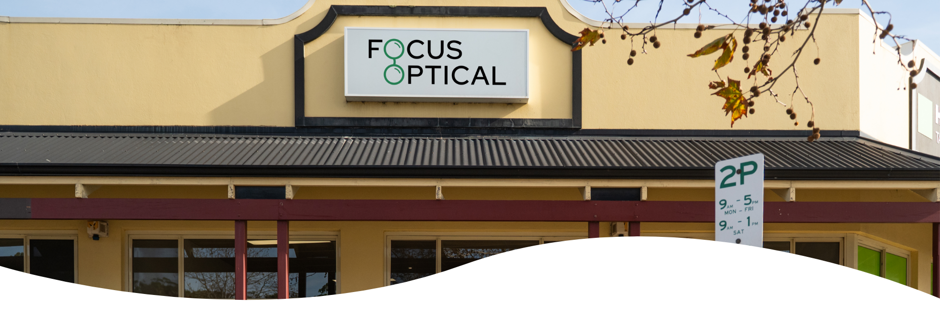 Facade of a building with a sign for Focus Optical, a parking sign indicating restricted parking hours, and tree branches with leaves and berries in the upper right corner.
