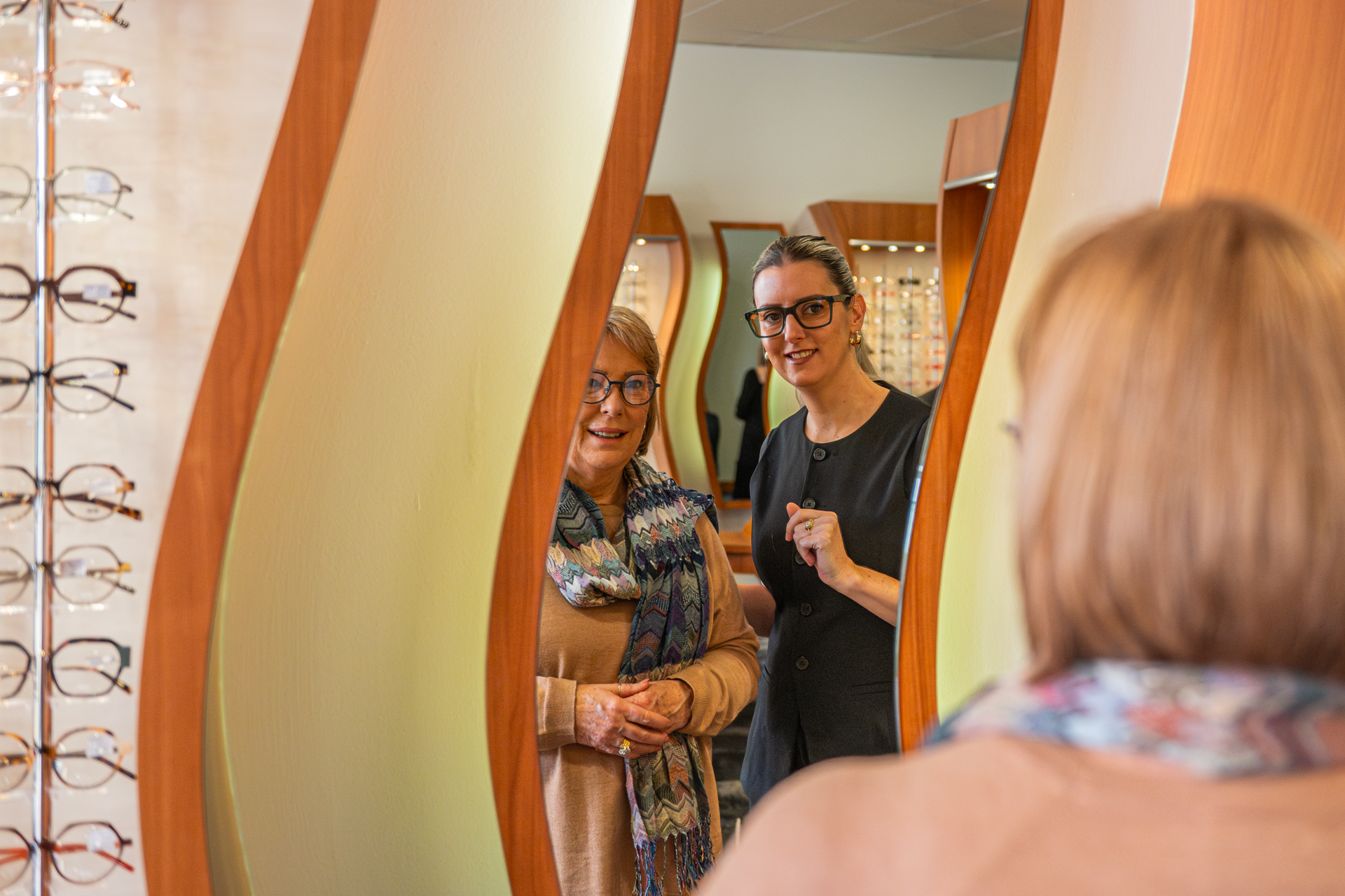 Two women looking at their reflection in a decorative mirror at an eyewear shop.