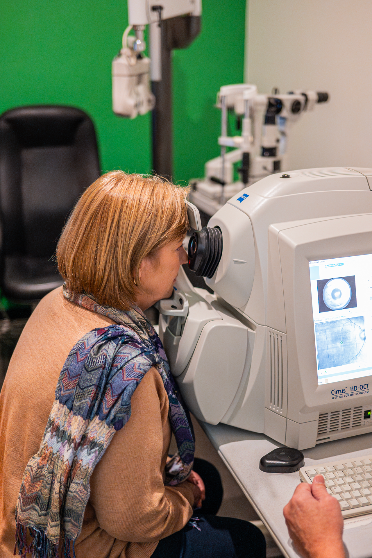 A woman with short auburn hair looking through a slit of a spectral domain optical coherence tomography (SD-OCT) machine during an eye exam.