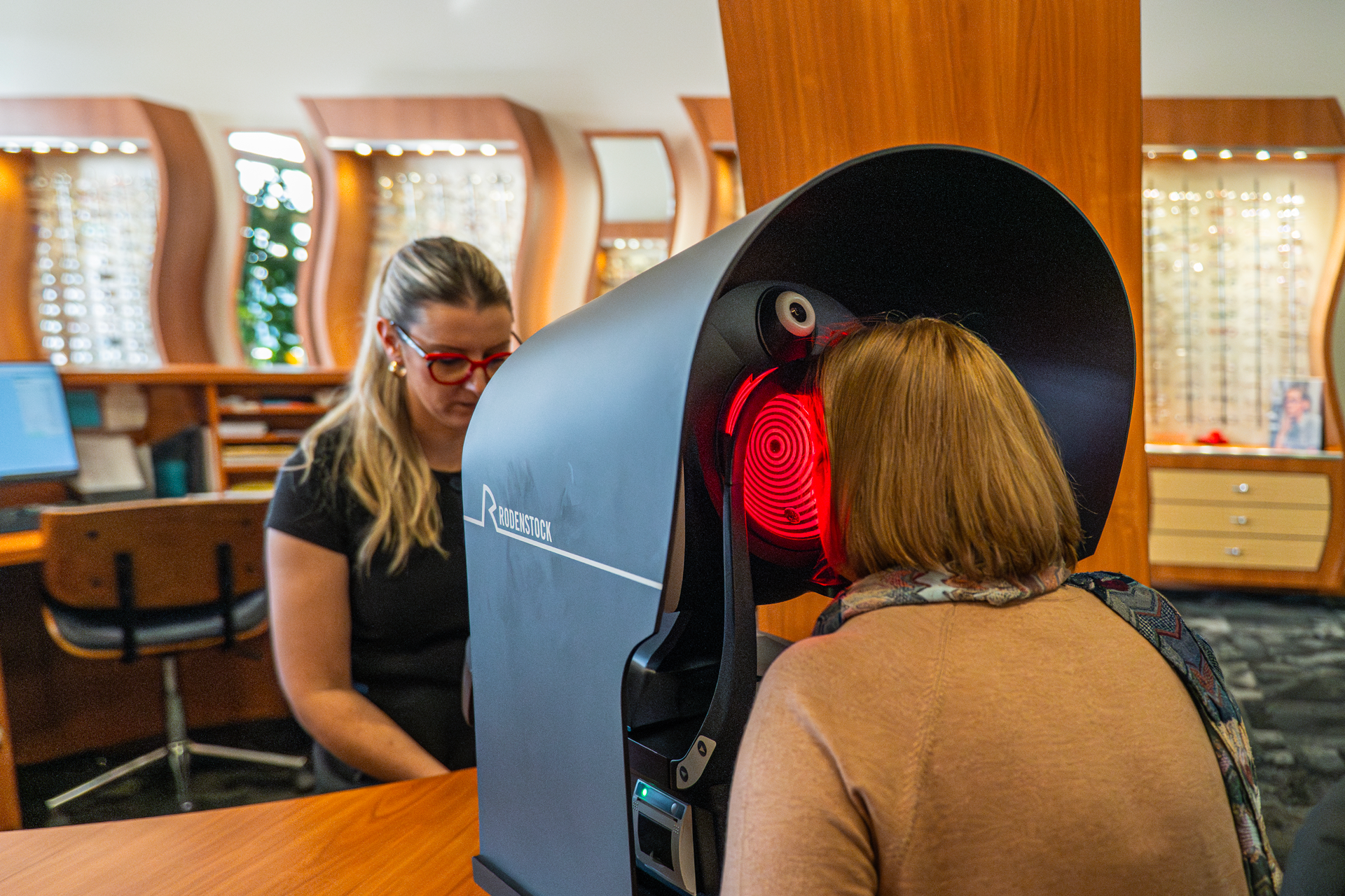 Woman with short brown hair using a retinal scanner, with a technician observing, in an optometry or vision testing office.