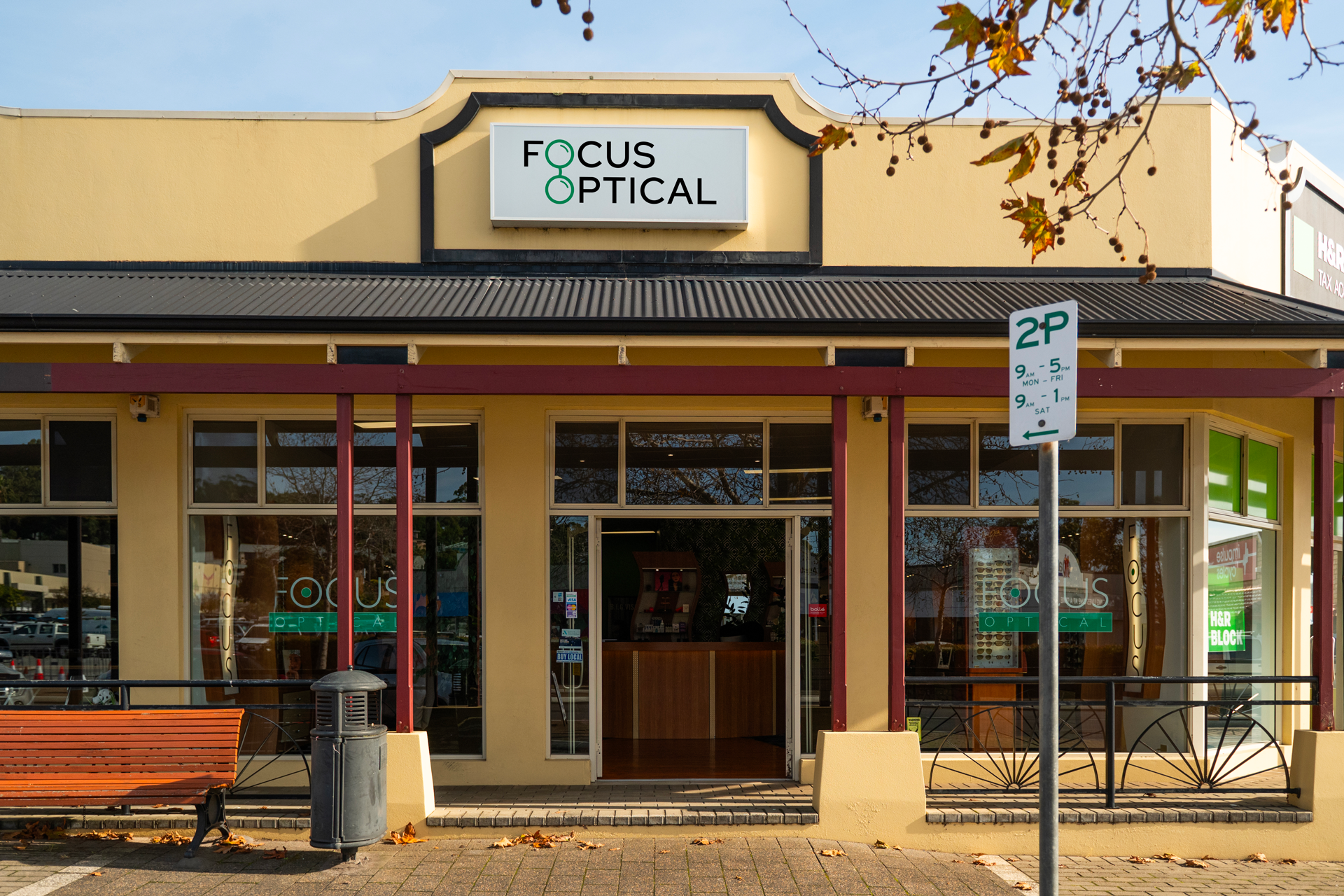 Exterior view of Focus Optical store with a yellow facade, red beams, large glass windows, a sign with the store's name, and a parking sign in front.