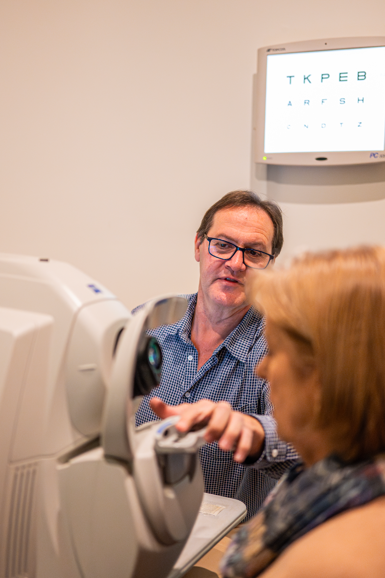 An optometrist conducts an eye exam for a woman using a phoropter, with an eye chart visible on a monitor in the background.