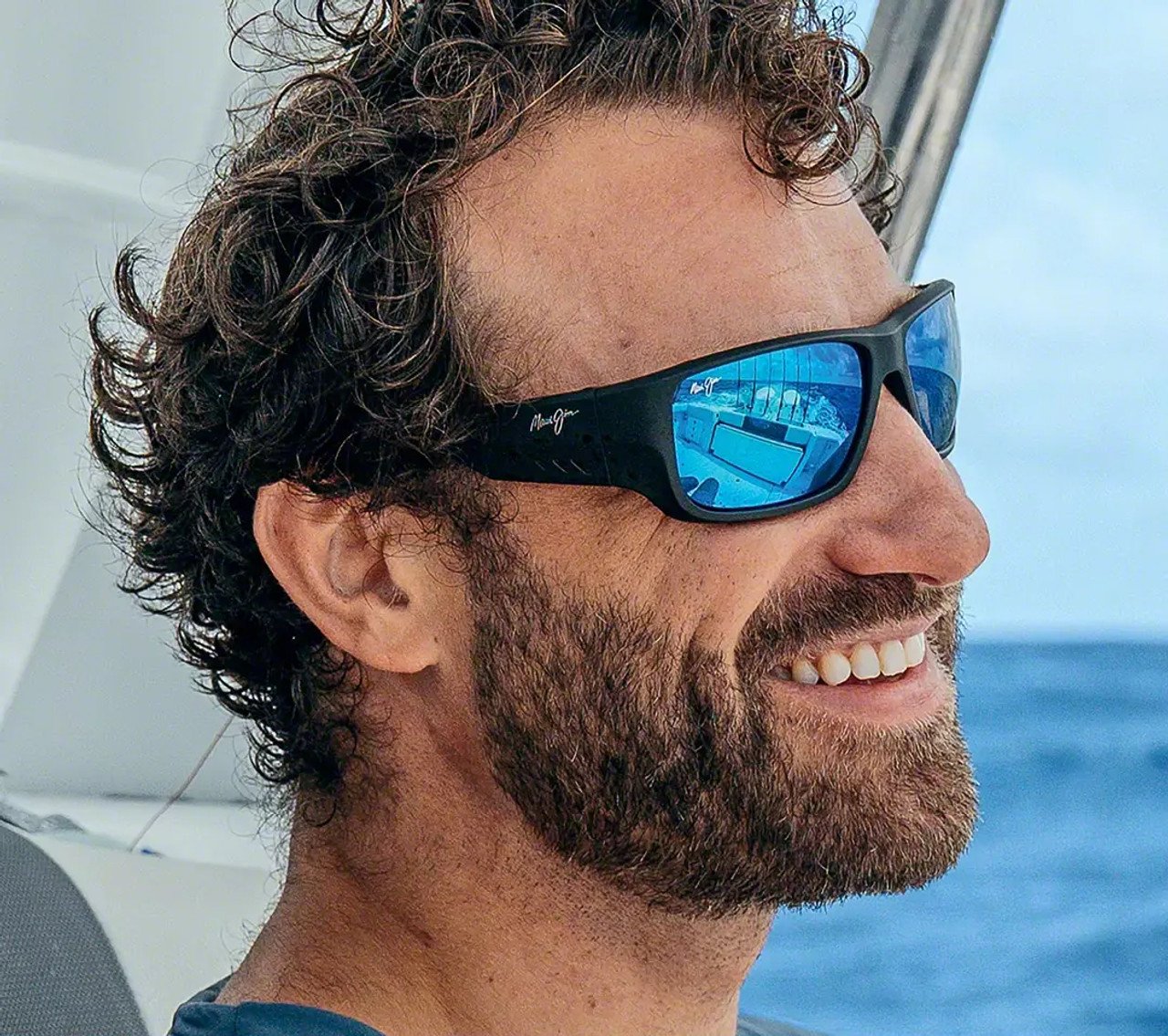 A man with curly hair and a beard wearing reflective sunglasses, smiling while on a boat with an ocean view in the background.