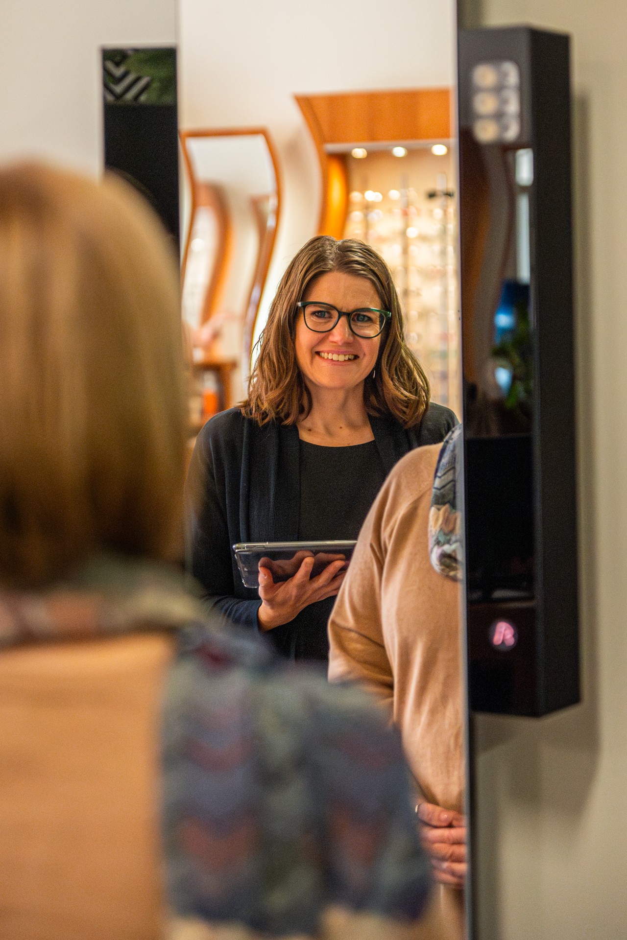 Woman with glasses looking into a mirror while holding a tablet, smiling, in a room with warm lighting.