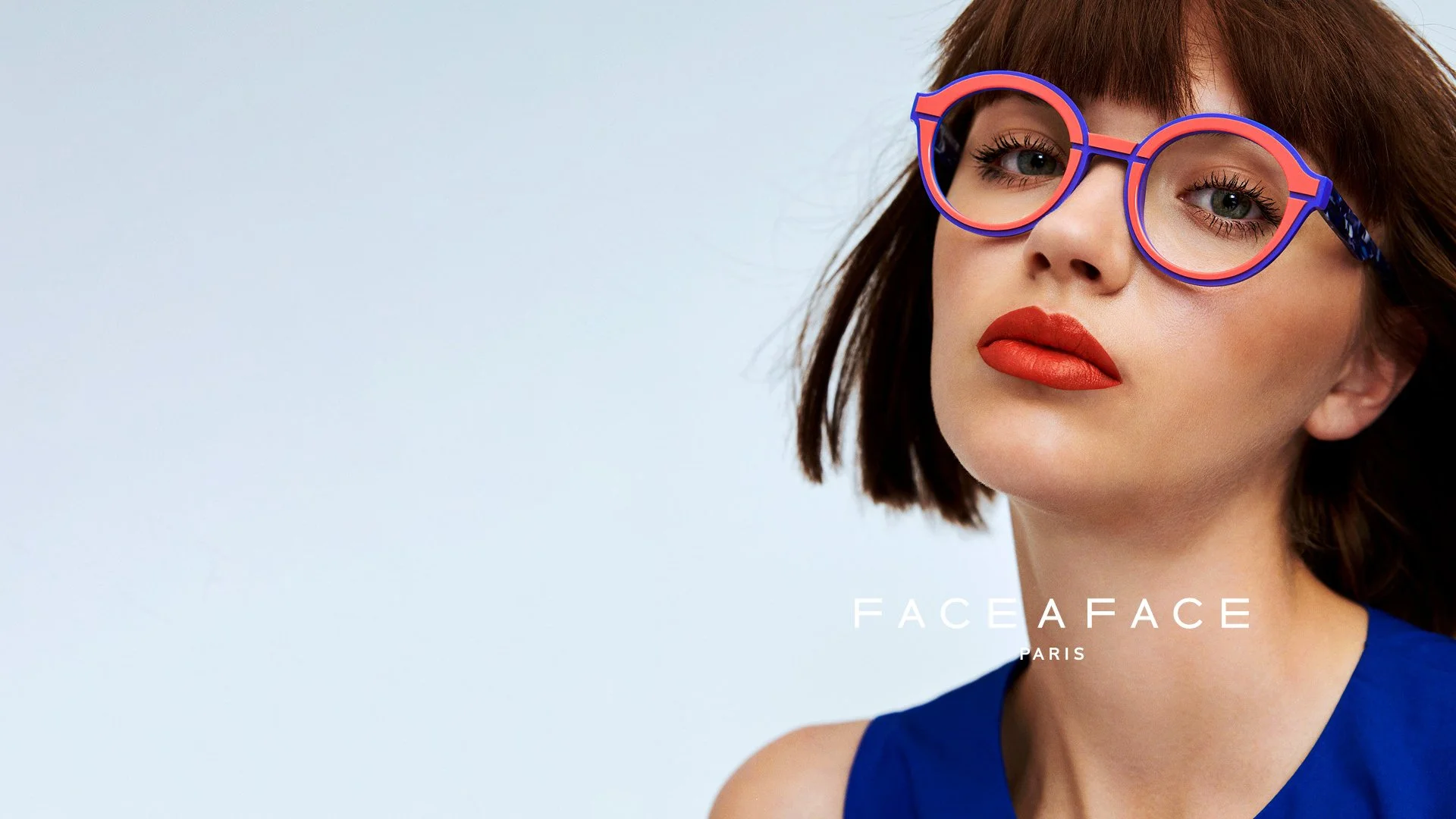 Close-up of a woman with red lipstick and glasses, wearing a blue sleeveless top, against a plain light background.