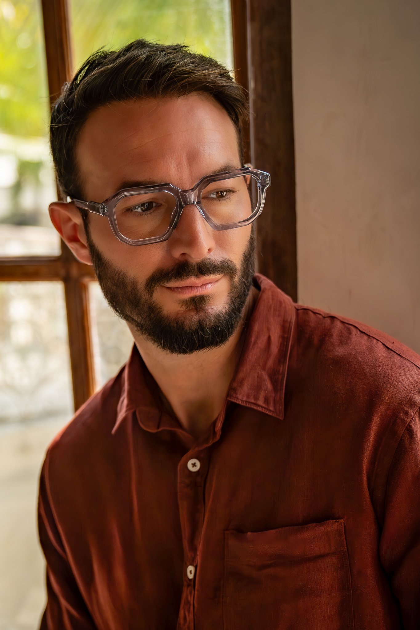A man with glasses and a beard looking out a window.