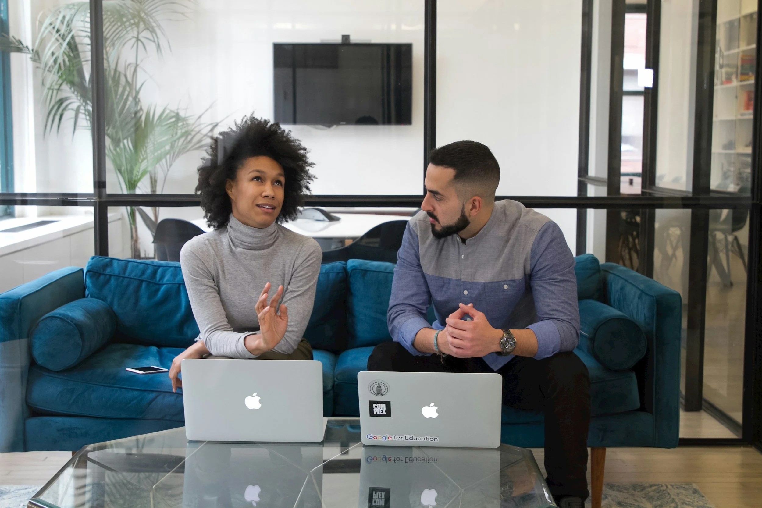 Two young adults, one woman and one man, having a conversation in an office lounge. They are sitting on a blue couch with laptops in front of them, and a glass coffee table with reflections. Behind them are large windows, a TV, a tall plant, and office furniture.