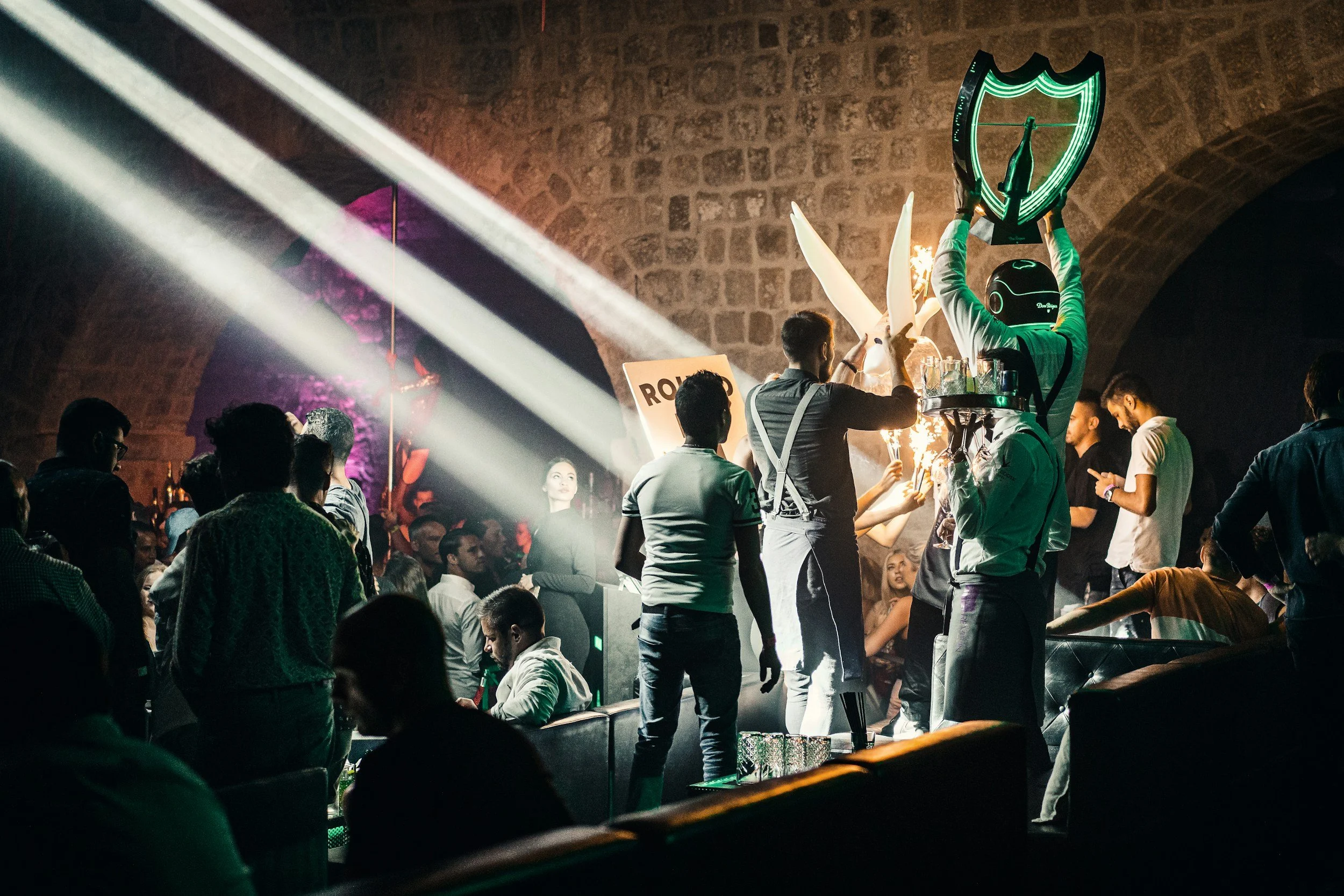 People gathered in a nightclub with a DJ wearing a helmet and holding a shield with a green emblem, surrounded by lights, sparklers, and a crowd enjoying the TikTok Live Creators party.