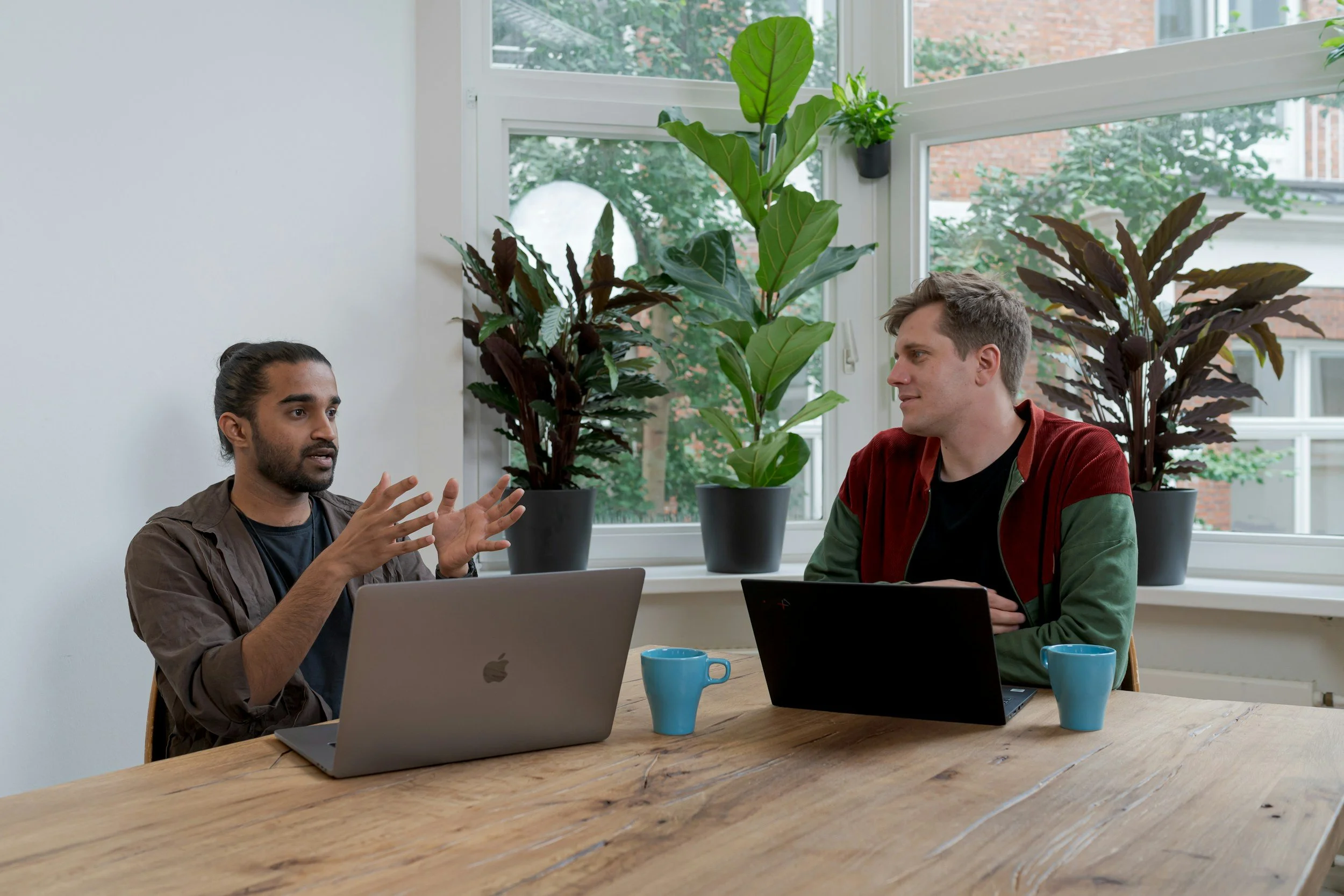 Two men having a discussion at a wooden table in a bright room with large windows and green plants.