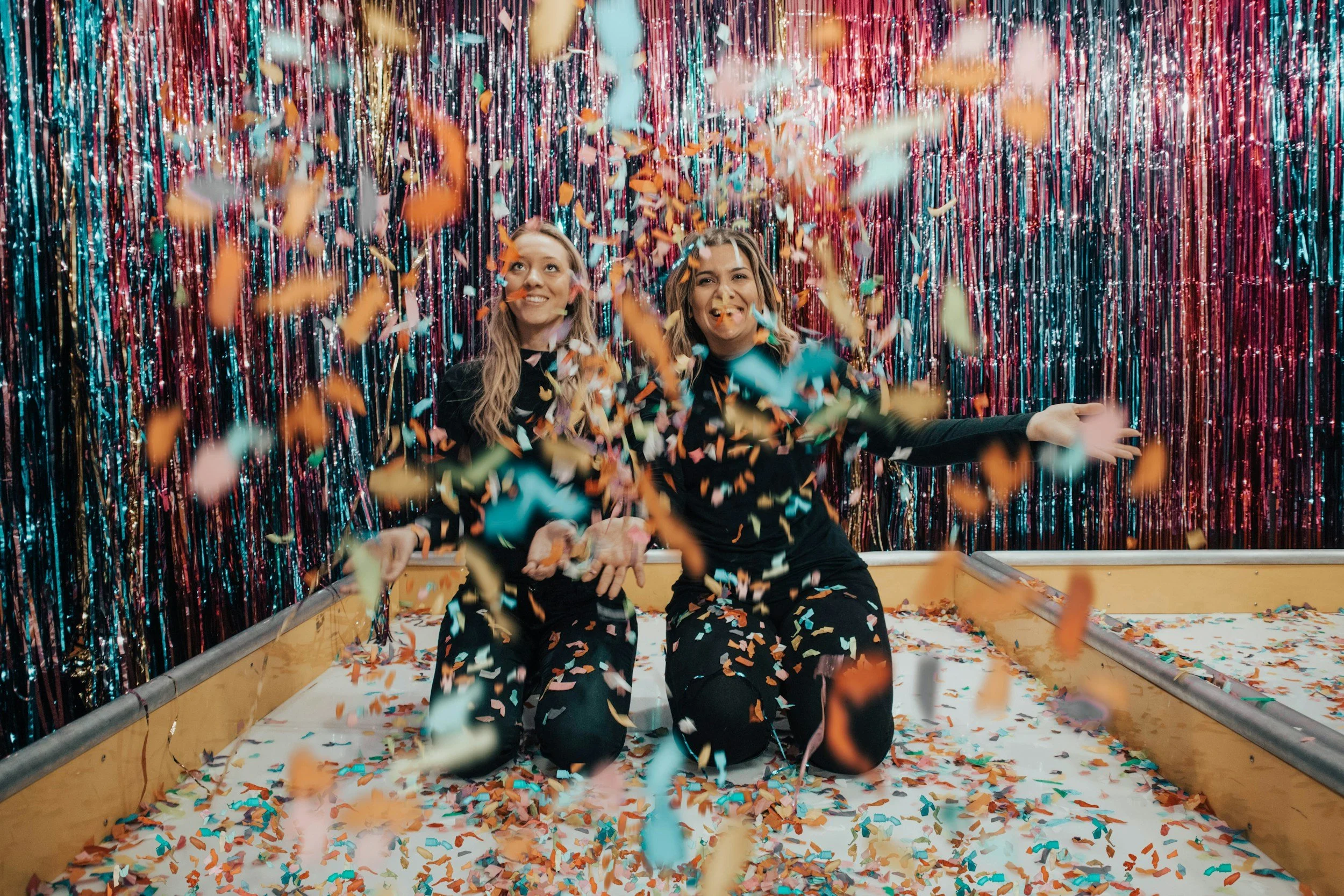 Two women in black clothing playing in a confetti-filled space with colorful metallic streamers as a backdrop.