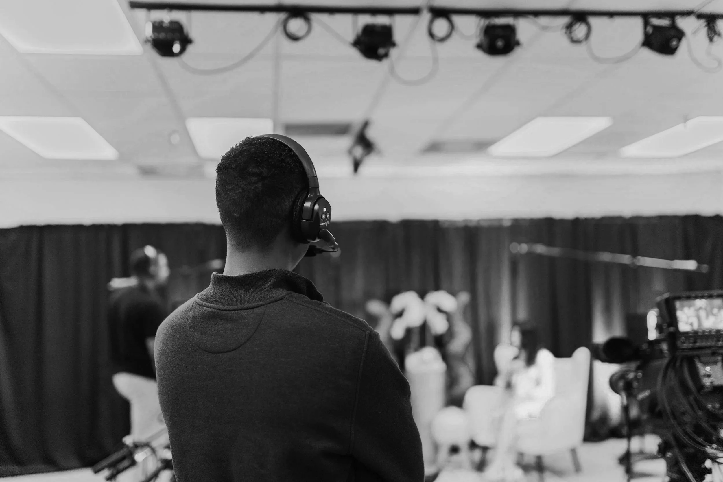 A person wearing a headset stands in a room with a camera set up, observing a stage with people and decorative swan figures in the background, likely behind the scenes at an event or recording.