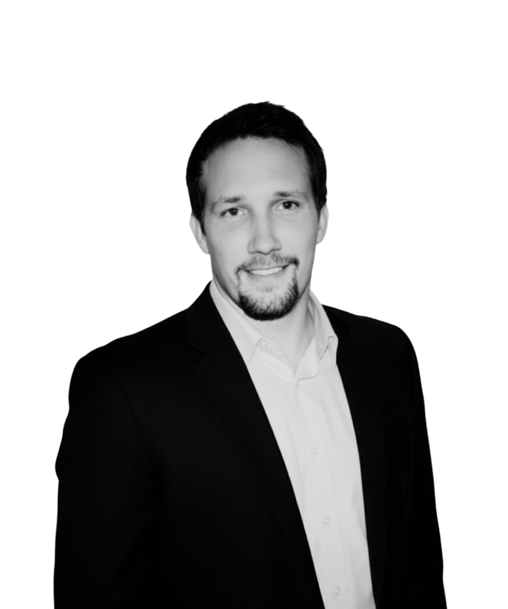 Black and white professional portrait of a man with short dark hair, a beard, wearing a suit and a white shirt, smiling slightly, against a black background.