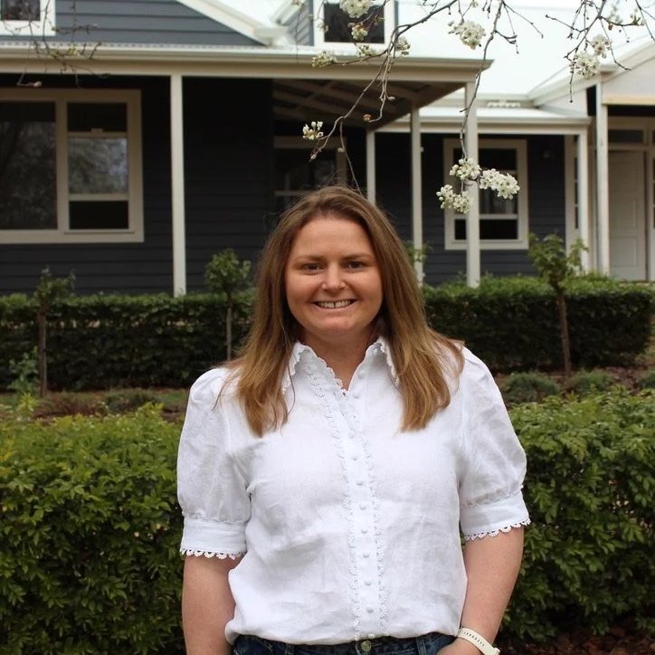 A smiling woman stands outdoors in front of a house with dark blue siding and white trim, surrounded by greenery and blooming white flowers.