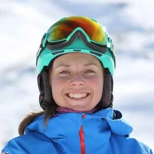 A woman wearing a teal ski helmet, orange ski goggles, and a blue ski jacket, smiling outdoors on a cloudy day.