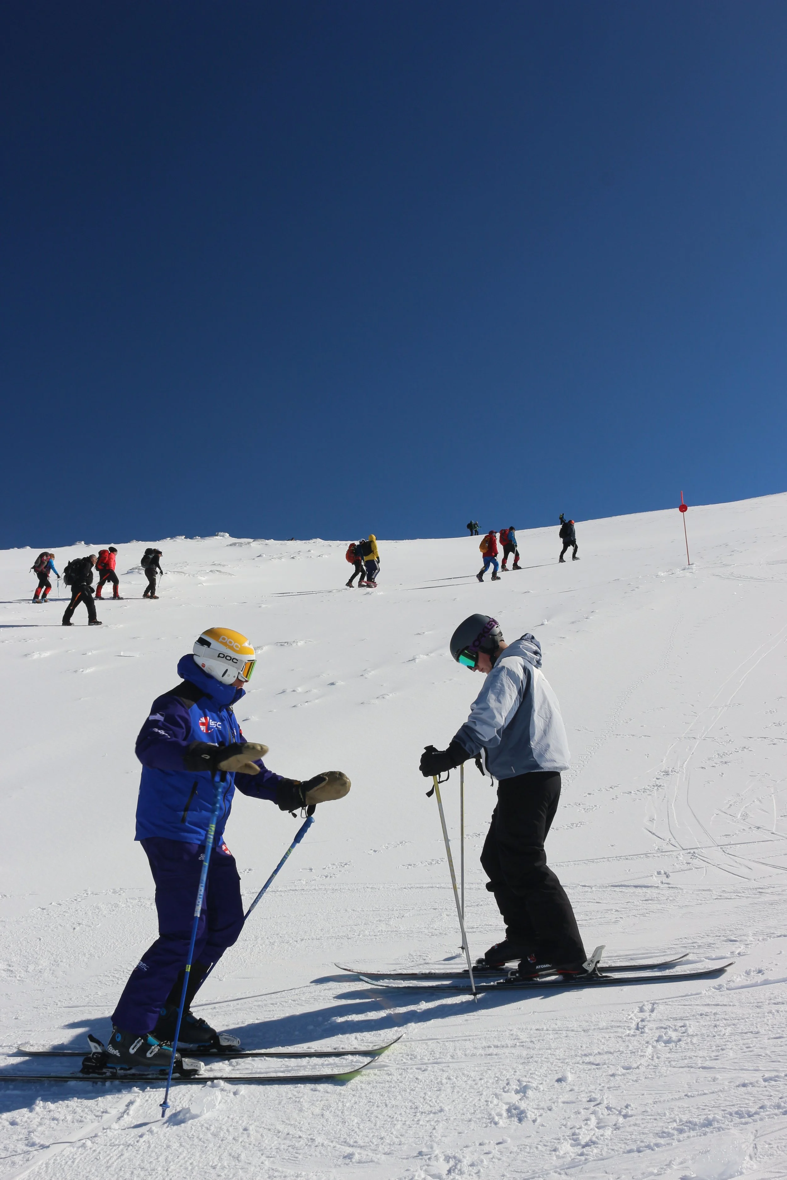 Two skiers on a snowy mountain slope, preparing to ski with several other skiers and snowboarders ascending the hill in the background under a clear blue sky.