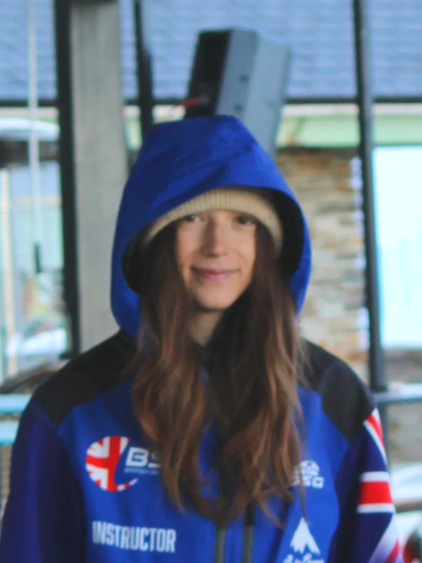Young woman in blue jacket with a hood, wearing a beige knit cap underneath, standing inside near large windows with brick and metal details.