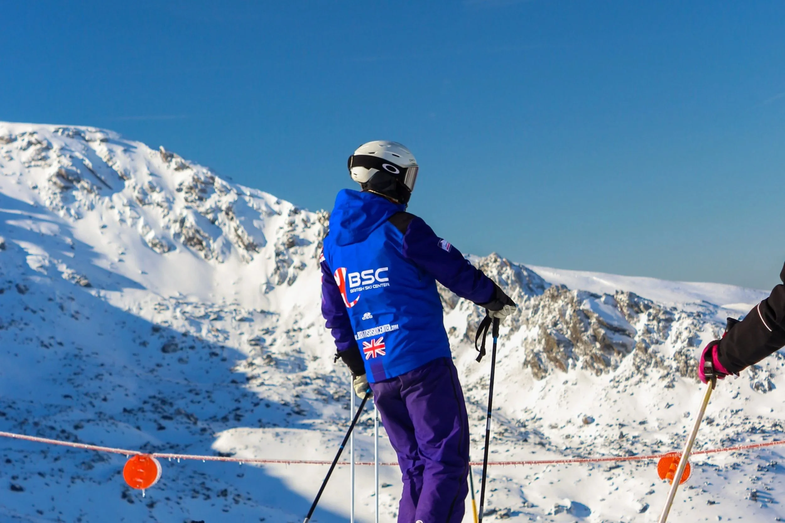 A person in a blue jacket and purple pants with a white helmet standing on snow in a mountainous area, holding ski poles, with snow-covered peaks in the background.