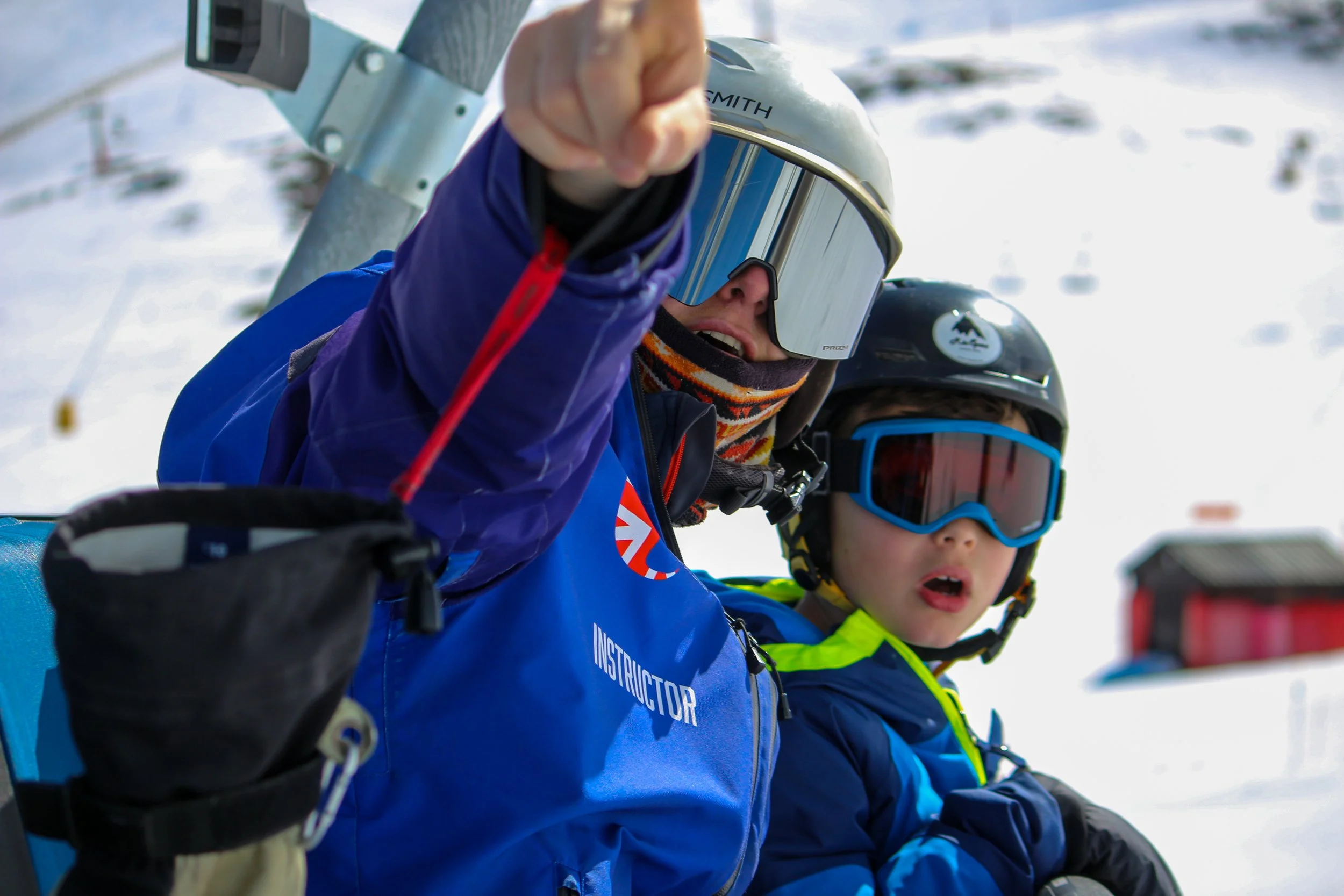 A ski instructor and a young student sitting on a ski lift in the snow, both wearing helmets and goggles, with the instructor pointing forward.