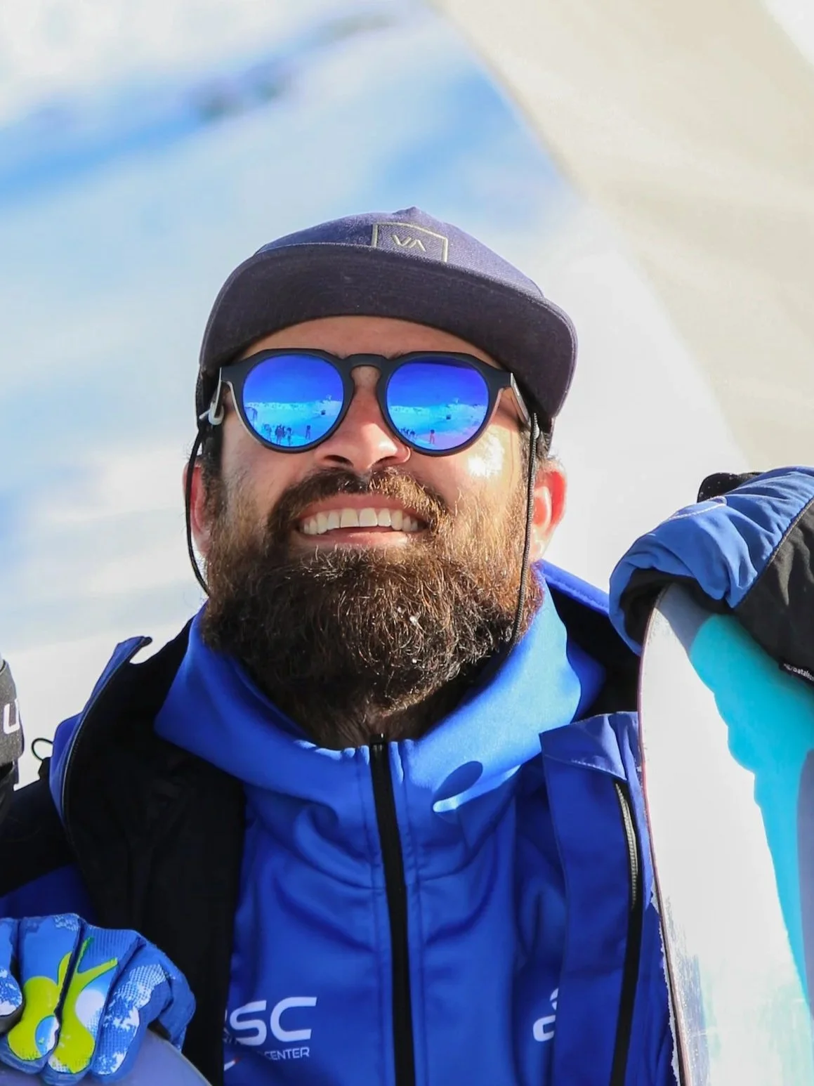 Man with beard wearing blue ski jacket, black sunglasses with blue reflective lenses, black cap, and gloves, smiling outdoors in snowy mountain setting.