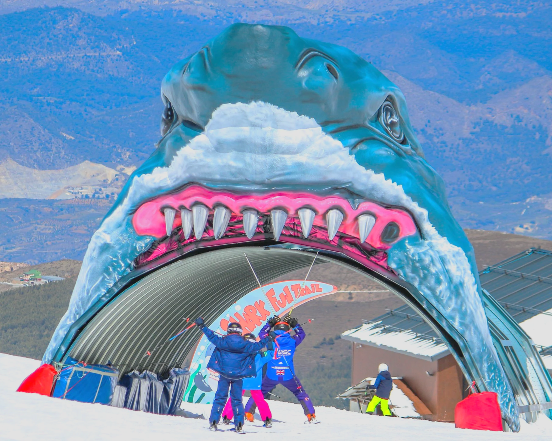 A large structure shaped like a shark head with sharp teeth, serving as an entrance for a ski trail, with children and adults in winter clothing playing and skiing in front of it.