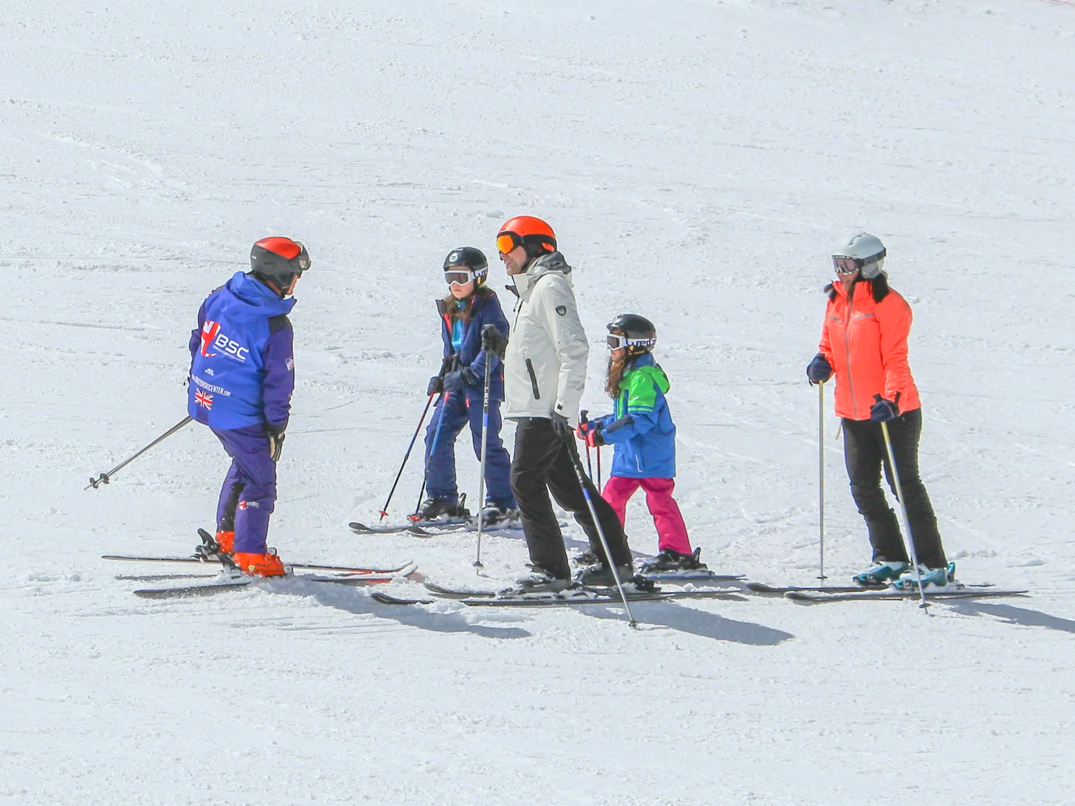 Group of five skiers, including children, standing on a snowy slope with ski gear, wearing helmets and colorful ski jackets.