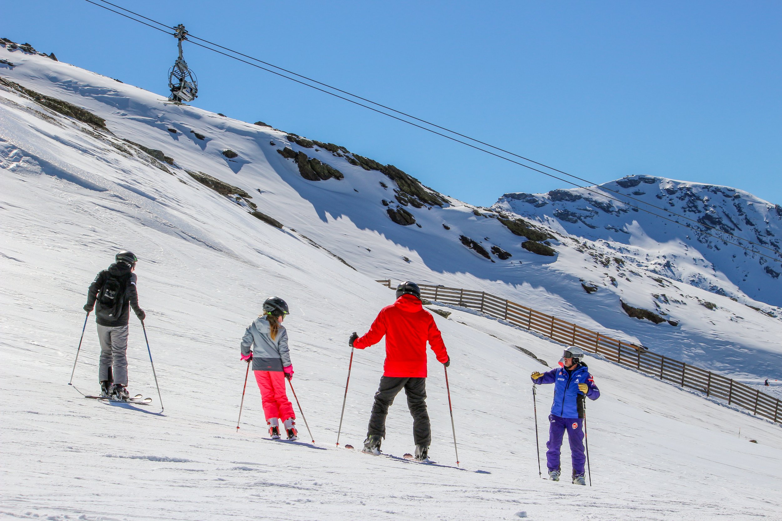 Group of five skiers in colorful winter gear skiing downhill on snow-covered mountain with ski lift and rocky peaks in background on clear, sunny day.