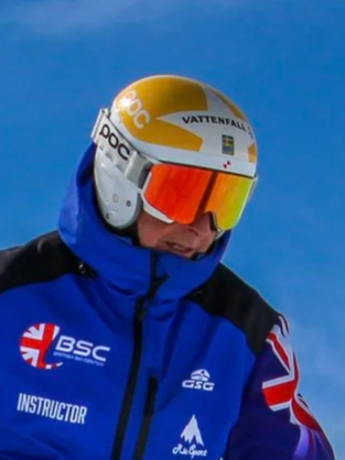 Close-up of a male ski instructor wearing a yellow helmet with logos and orange ski goggles, dressed in a blue jacket with patches and flags, against a clear blue sky.