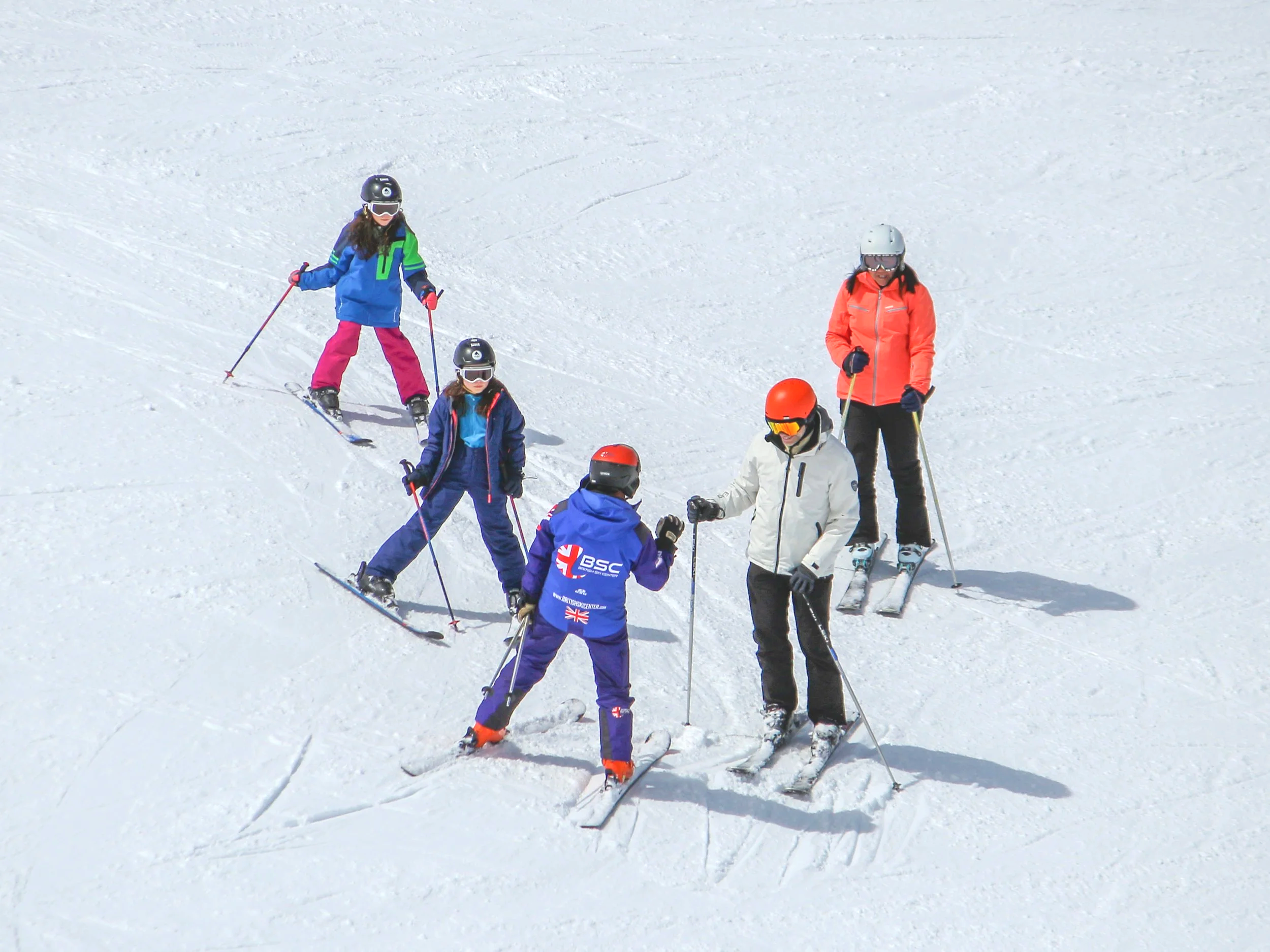 Group of six people skiing on a snow-covered slope, wearing colorful winter clothing and helmets.