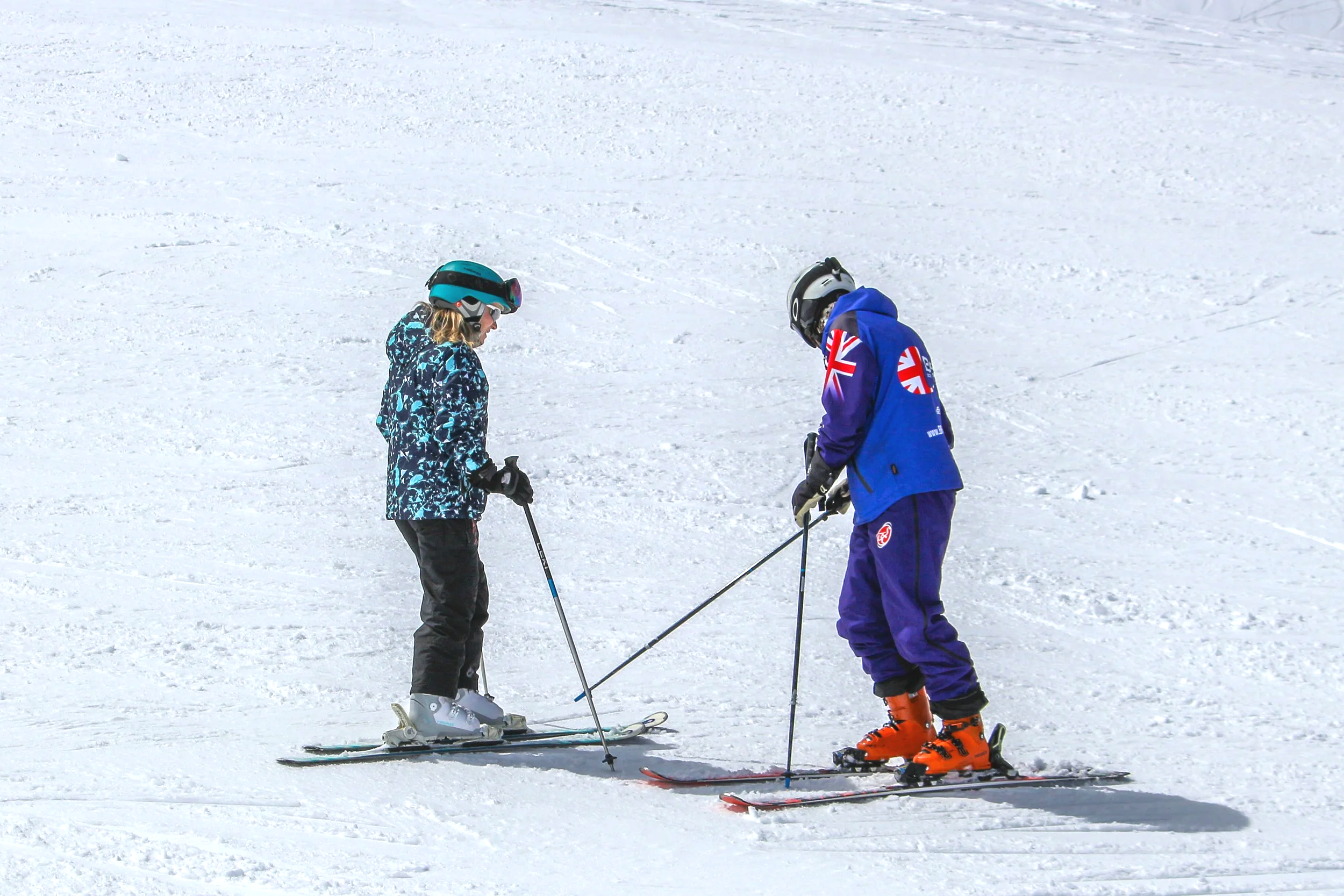 Two children in ski gear on a snowy slope, one wearing a blue patterned jacket and the other in a blue tracksuit with British flags, standing on skis with ski poles.