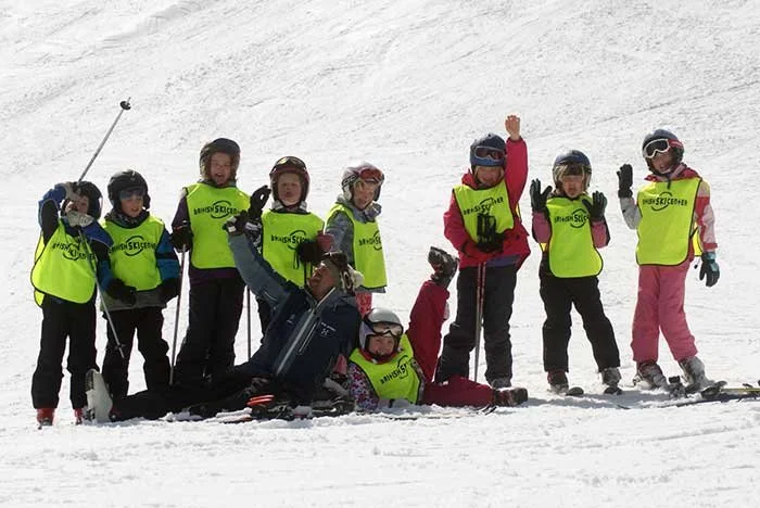 Group of children in ski gear posing on a snowy slope, some standing and some lying down, wearing bright yellow vests and helmets.