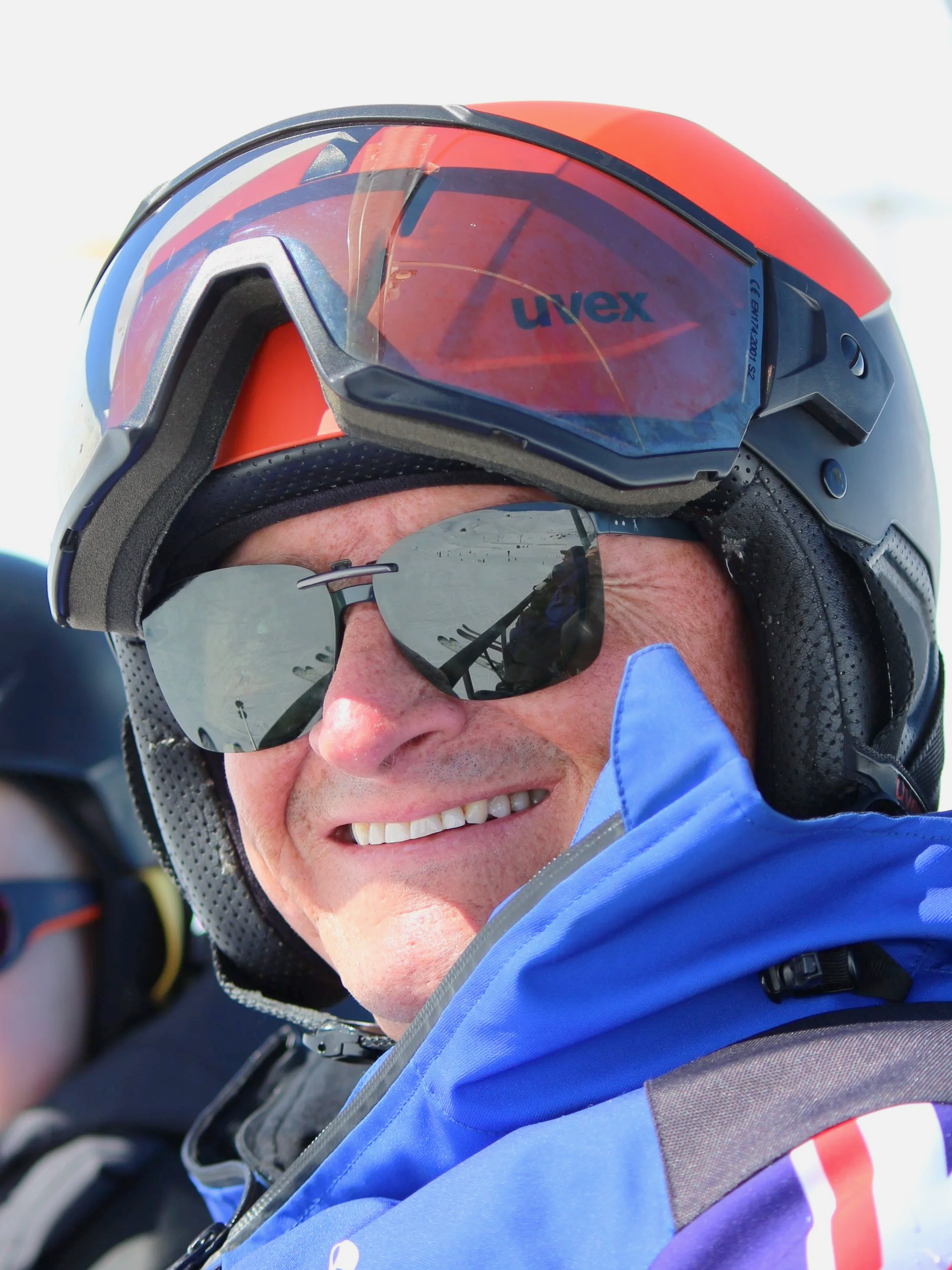 A smiling man wearing a black helmet with a red and black visor, sunglasses, and a blue jacket, likely outdoors.