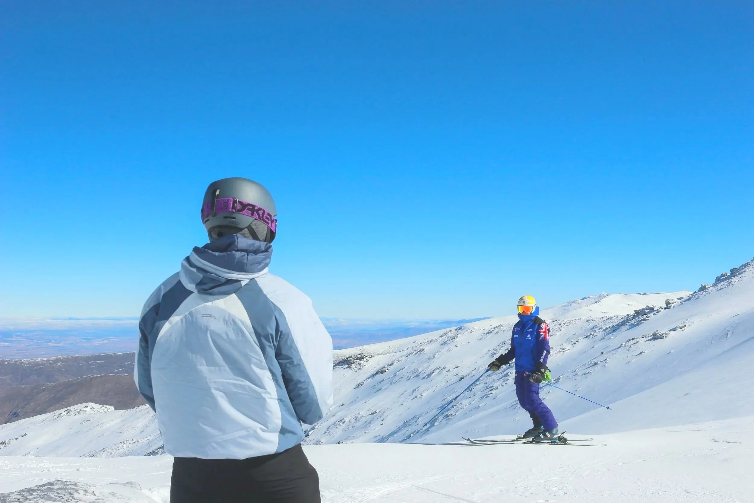 Two skiers on a snowy mountain slope with clear blue sky and distant mountains in the background.