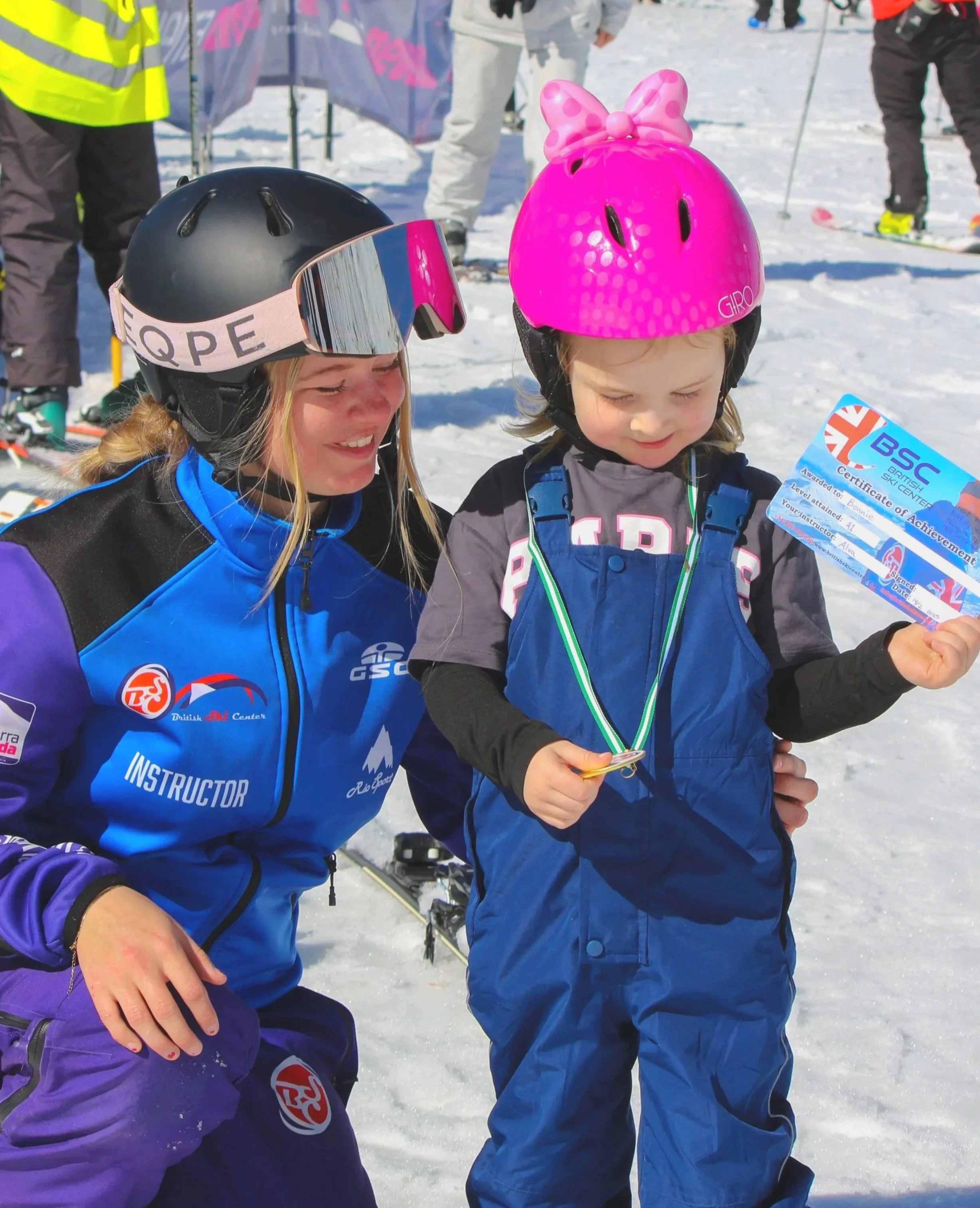 A young girl wearing a pink helmet with a bow and a blue ski suit, holding a certificate and a medal, is standing on a snowy slope with an adult instructor smiling nearby.