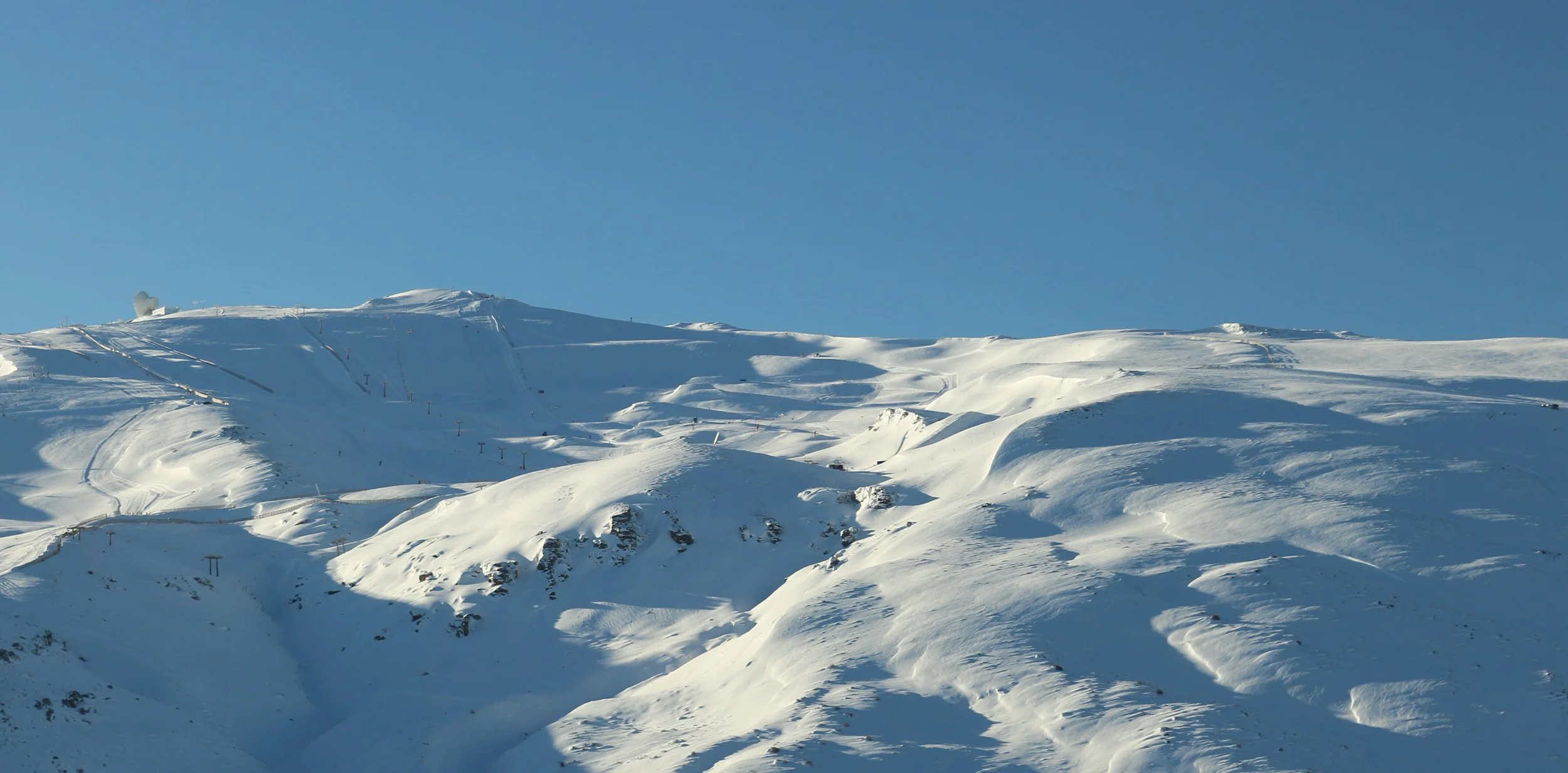 Snow-covered mountain slope under a clear blue sky with ski lifts and a satellite dish at the top.