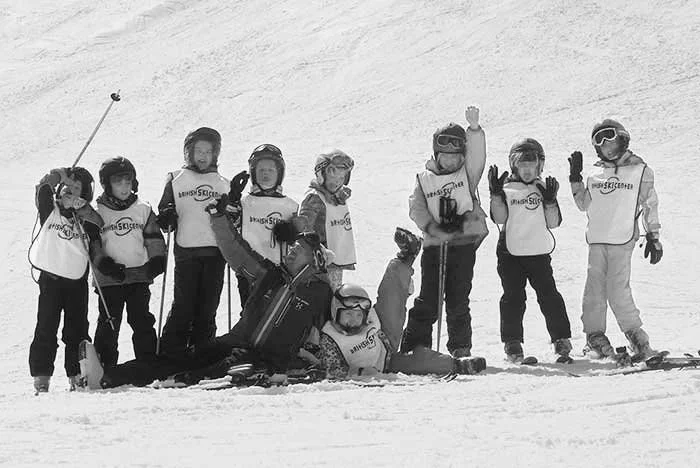 Group of children in ski gear and helmets posing on snowy slope, some making peace signs, one sitting with skis, others standing with ski poles.